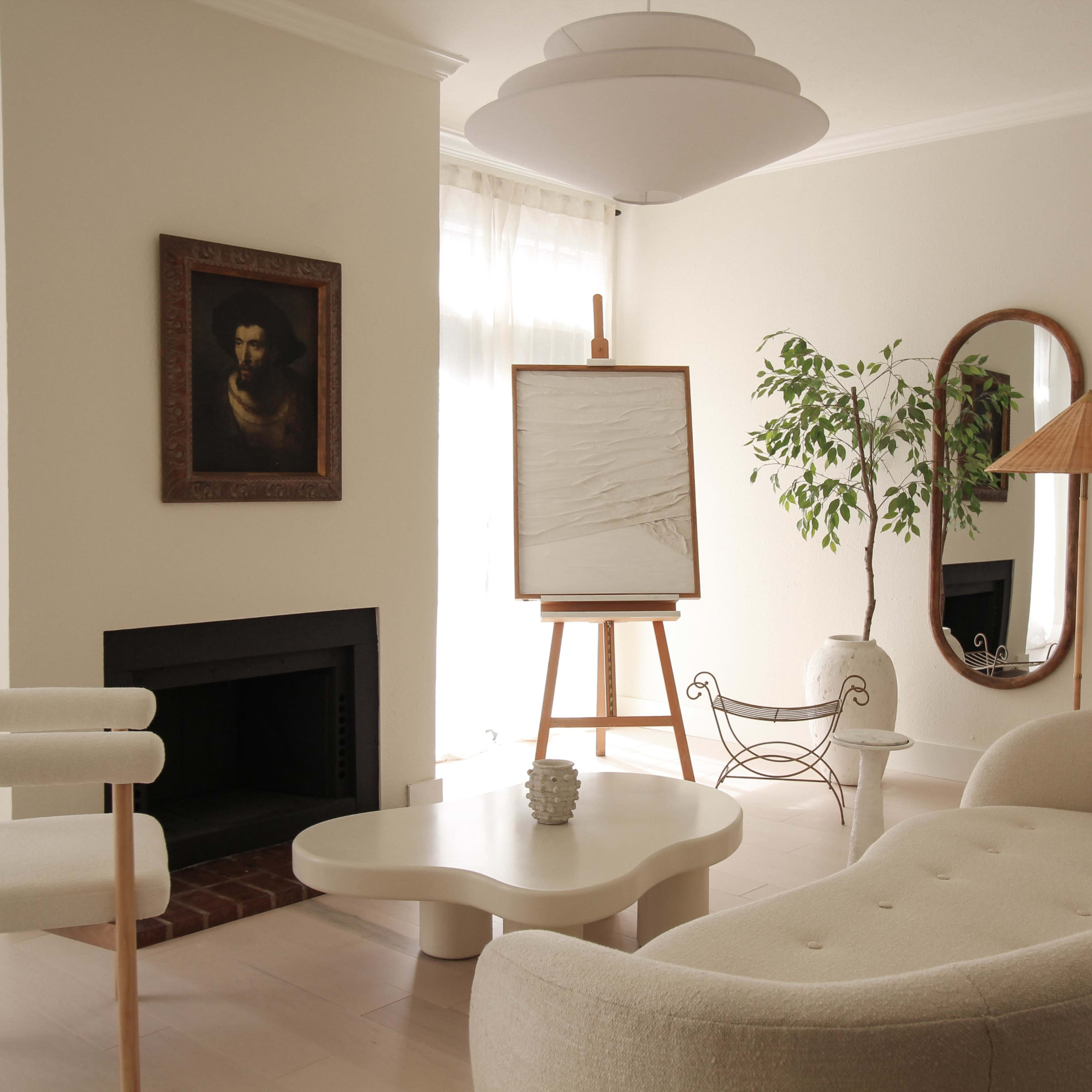 A modern living room featuring a light-colored sofa, a minimalistic coffee table, an easel with a blank canvas, and a large indoor plant.