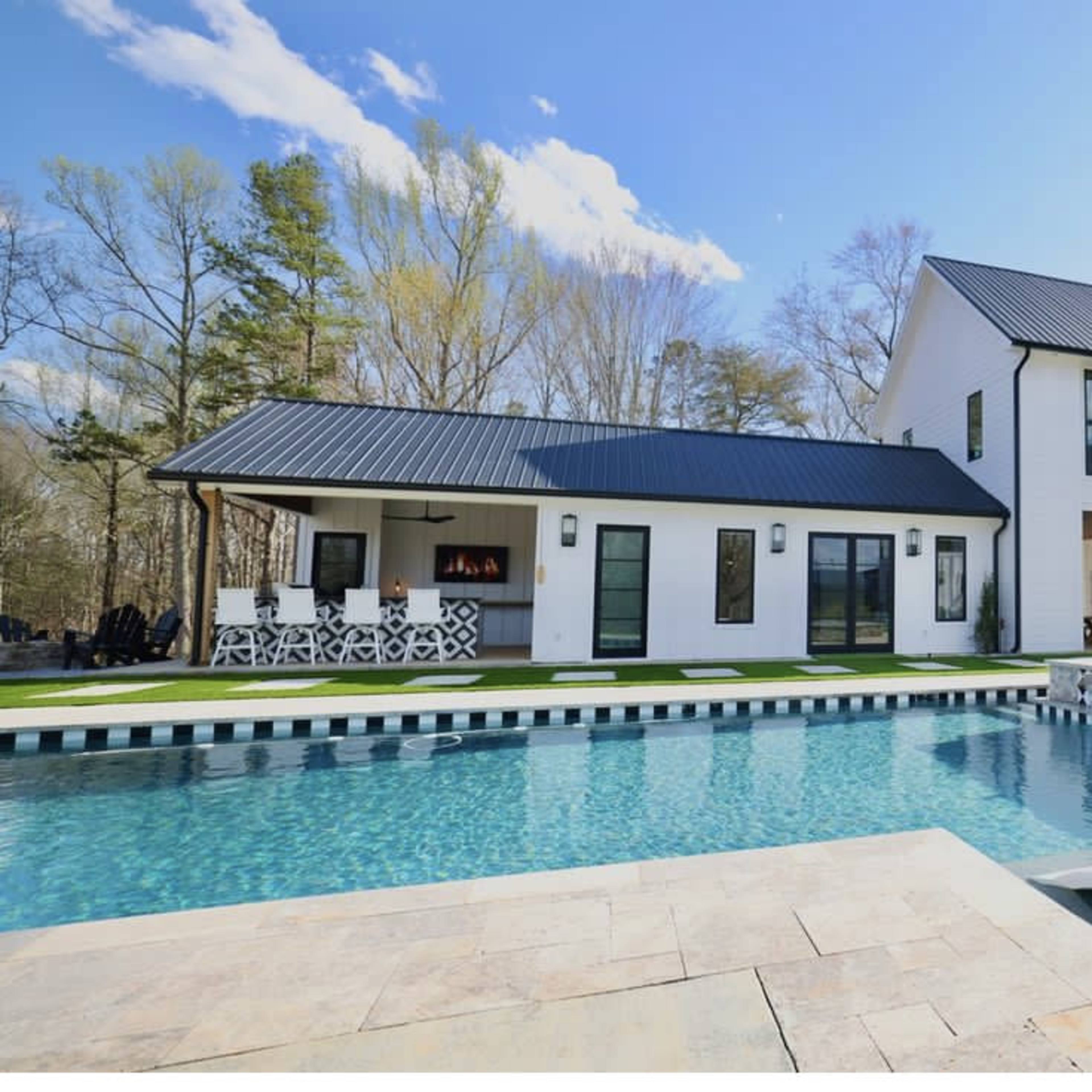 A modern two-story house with large windows and a black metal roof is situated beside a swimming pool featuring a spa and lounge chairs.