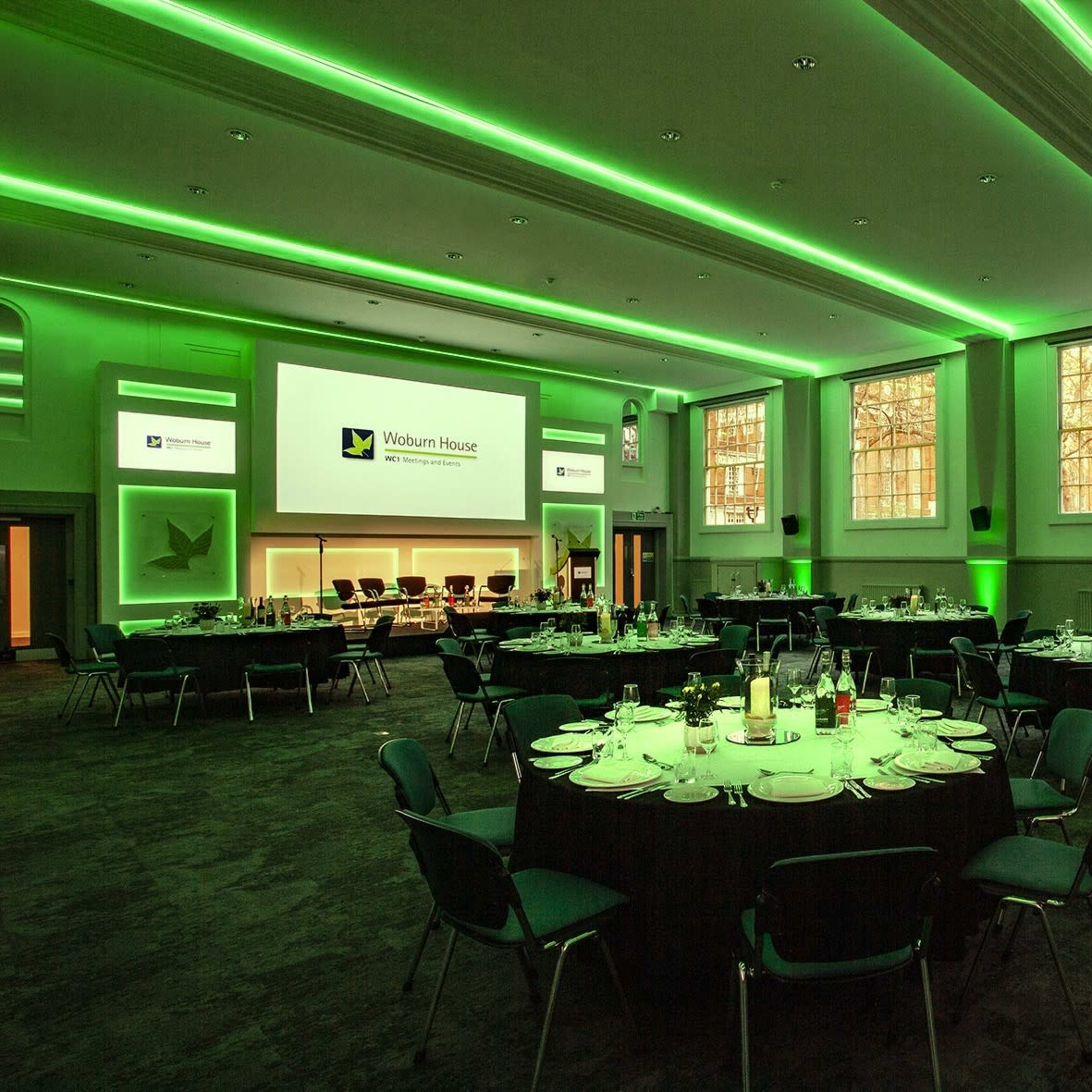 A banquet hall featuring round tables set for dining, illuminated with green lighting and equipped with a large screen for presentations.