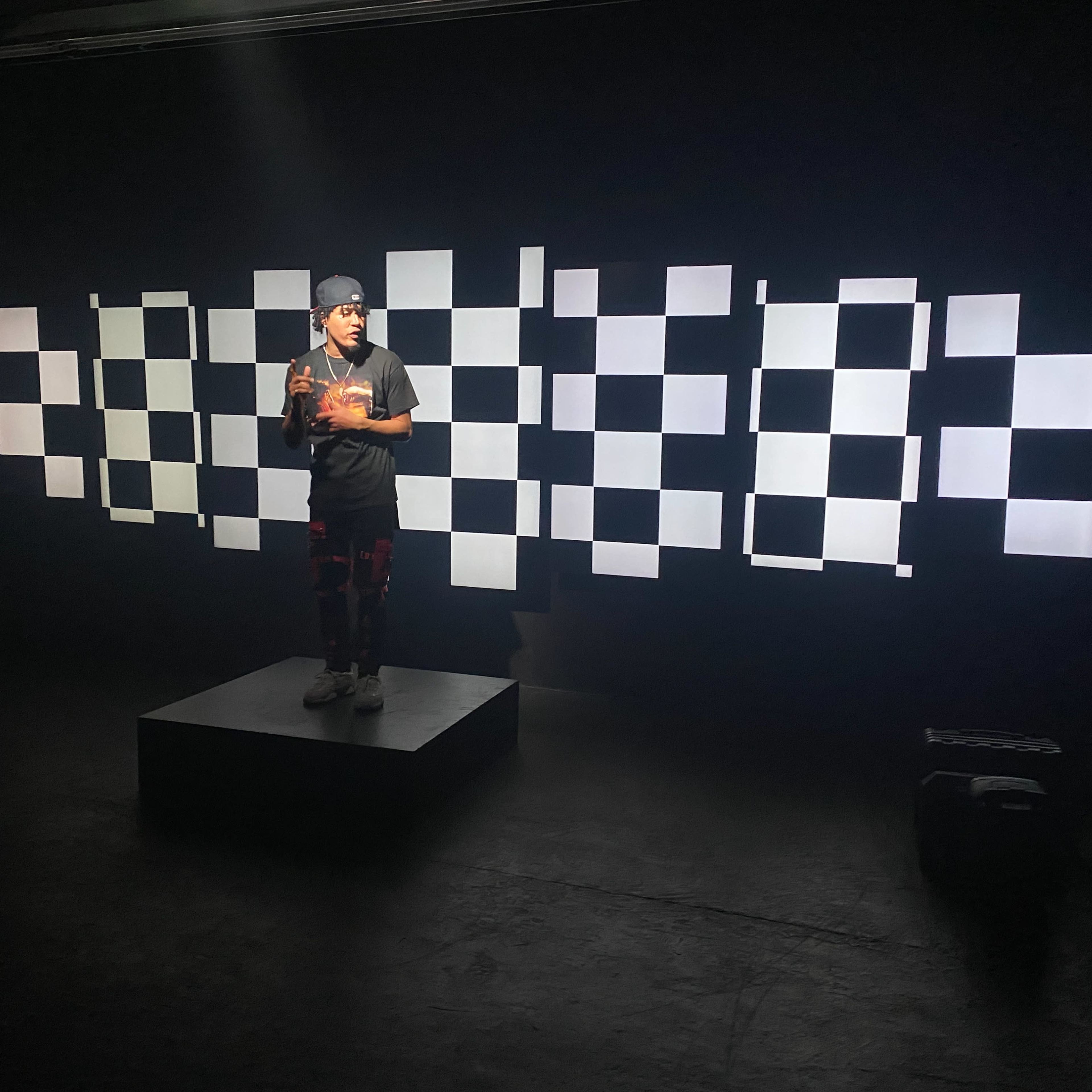 A person stands on a platform in a dark room, surrounded by a backdrop of alternating light and dark squares.