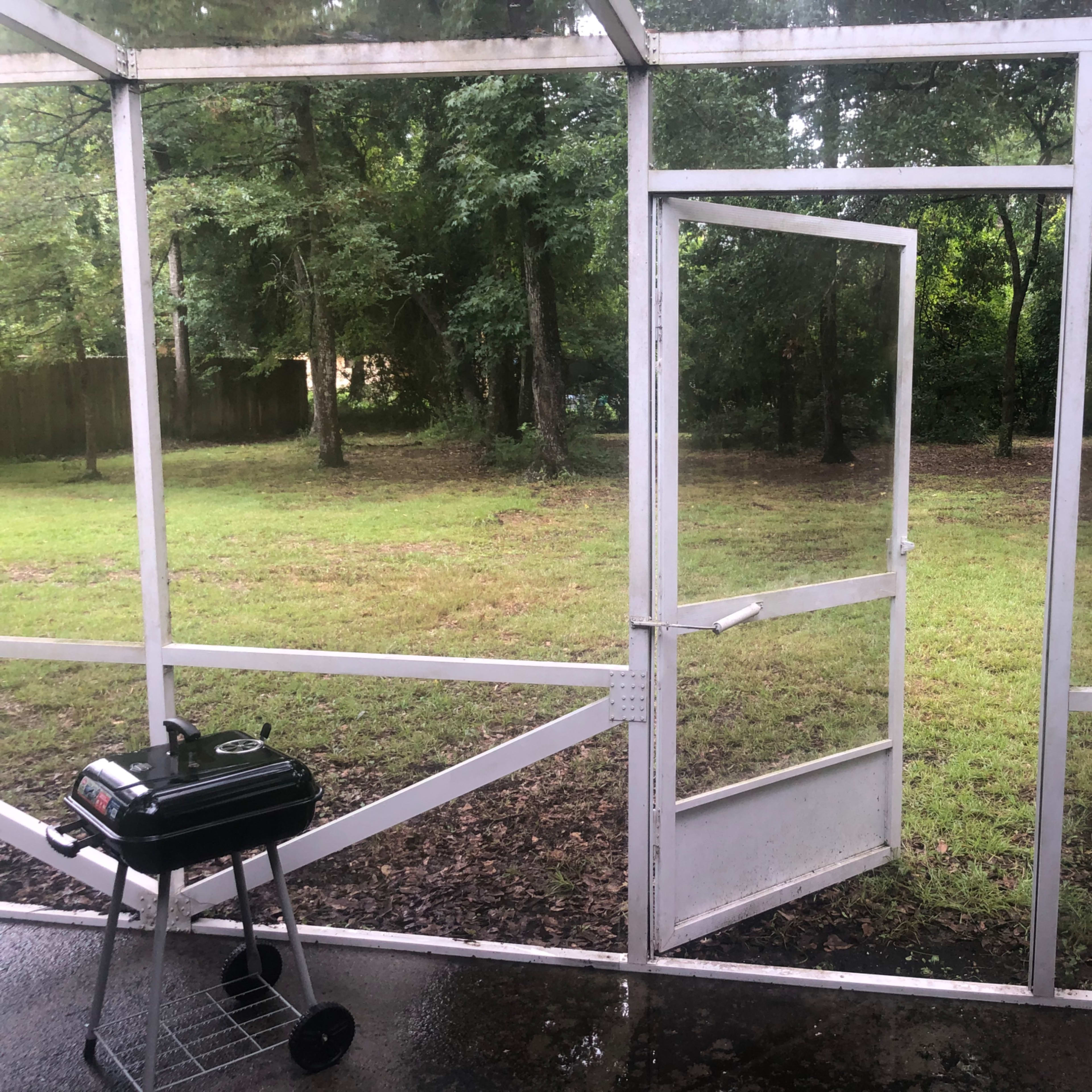 An open screen door leading to a grassy area with trees, alongside a small charcoal grill on the porch.