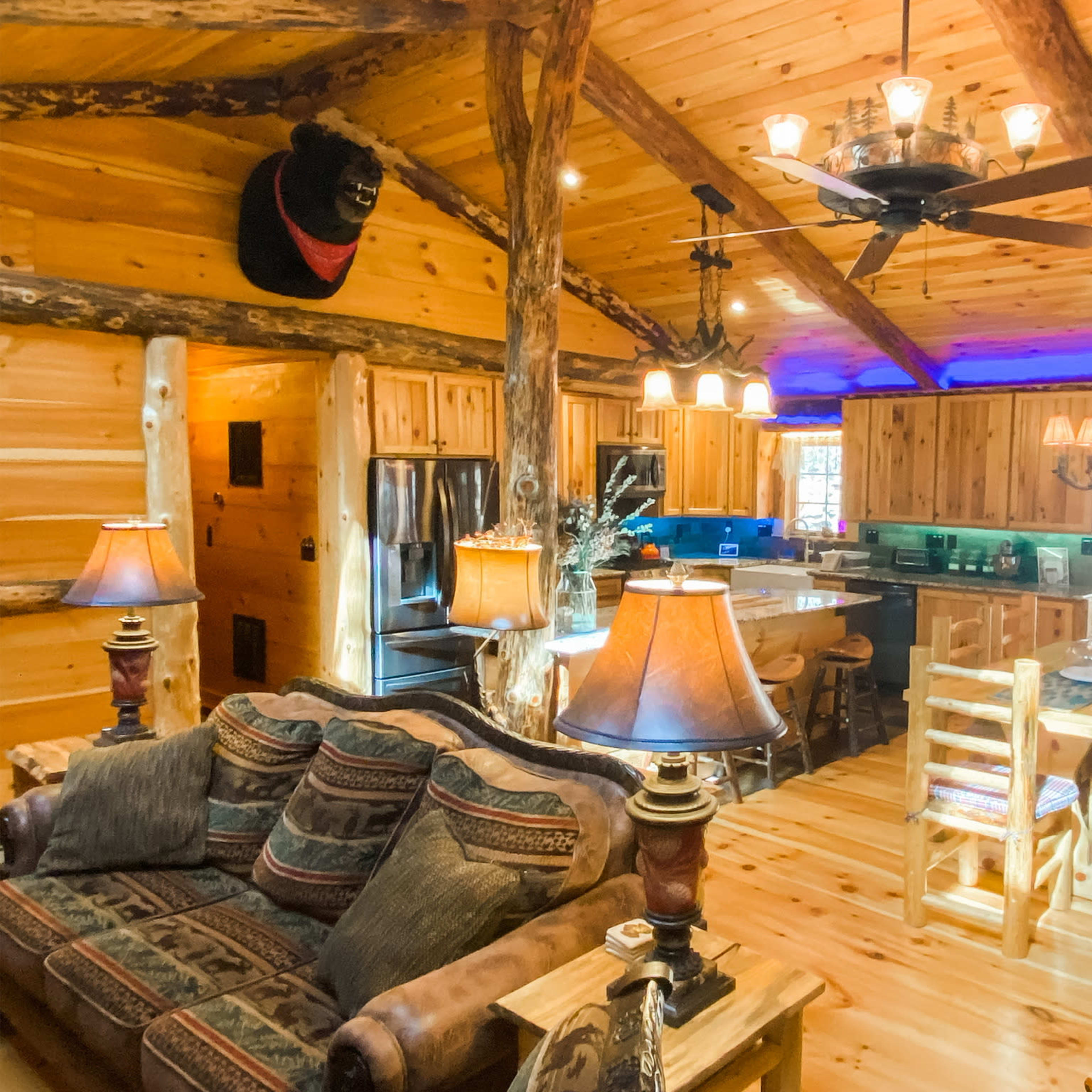A rustic wooden cabin interior with a living area featuring a couch, a dining table, and a kitchen area in the background.