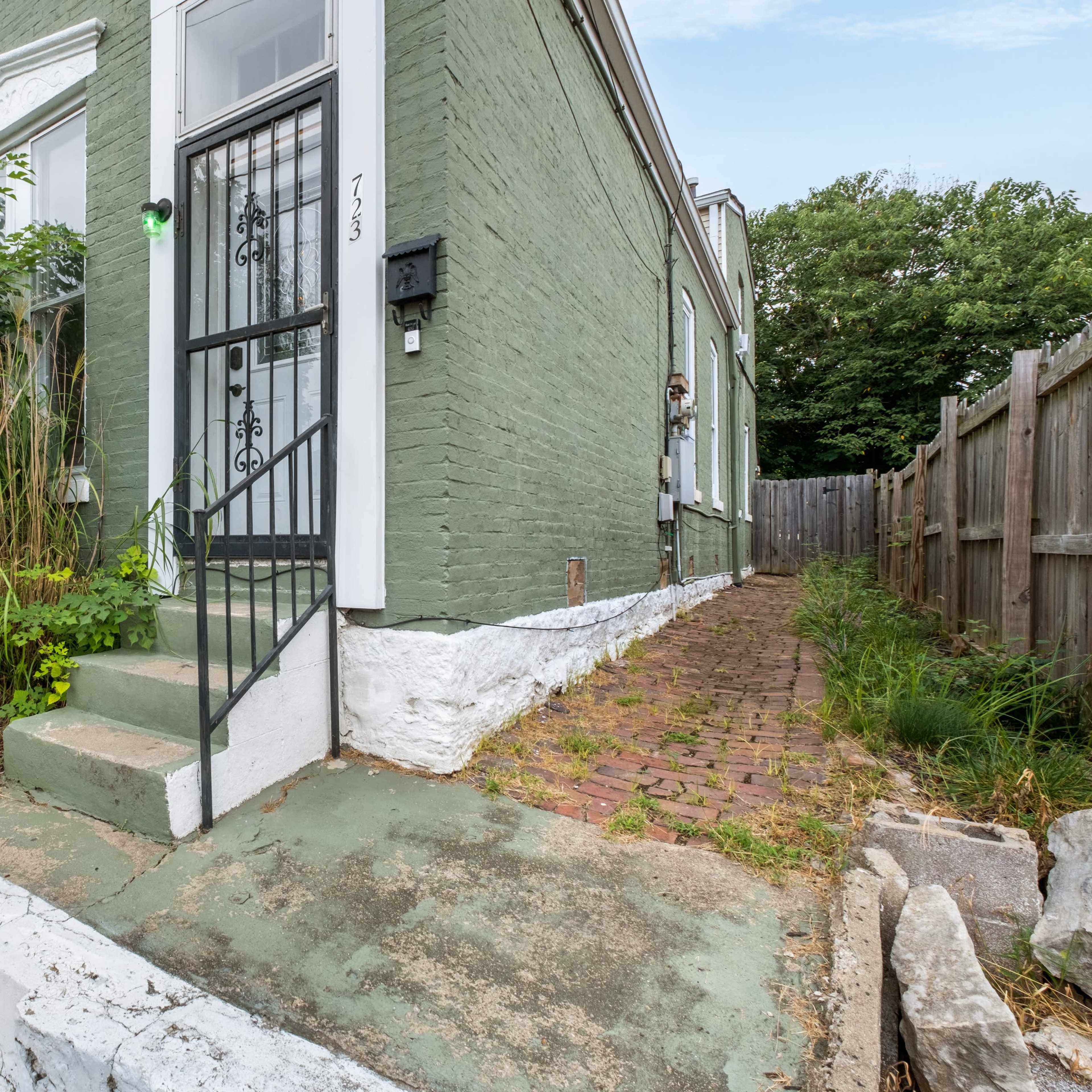 A green-painted house with a staircase leading to a front door, surrounded by overgrown plants and a brick pathway beside a wooden fence.