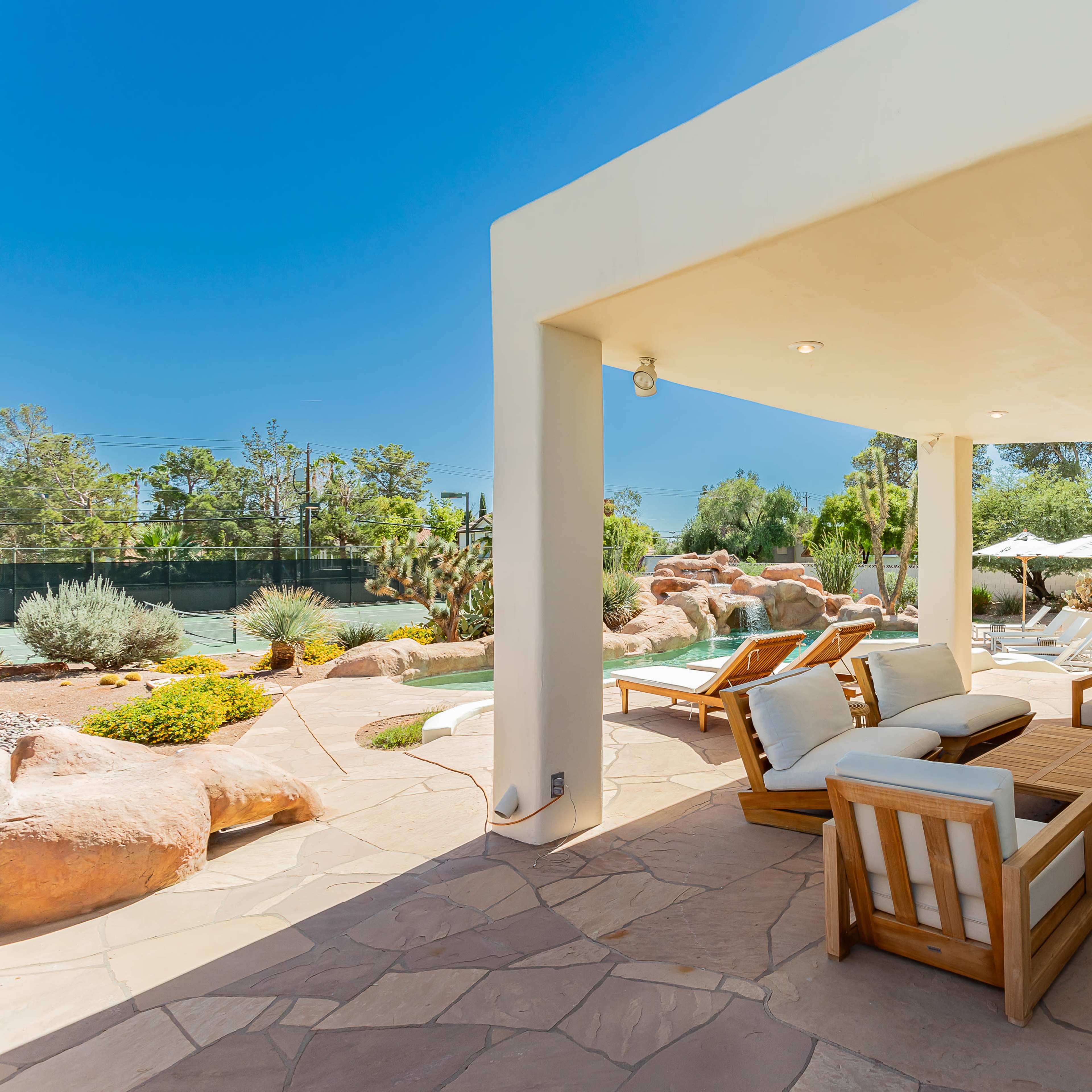 A modern outdoor seating area features wooden furniture and a stone patio, surrounded by lush greenery and rock formations.