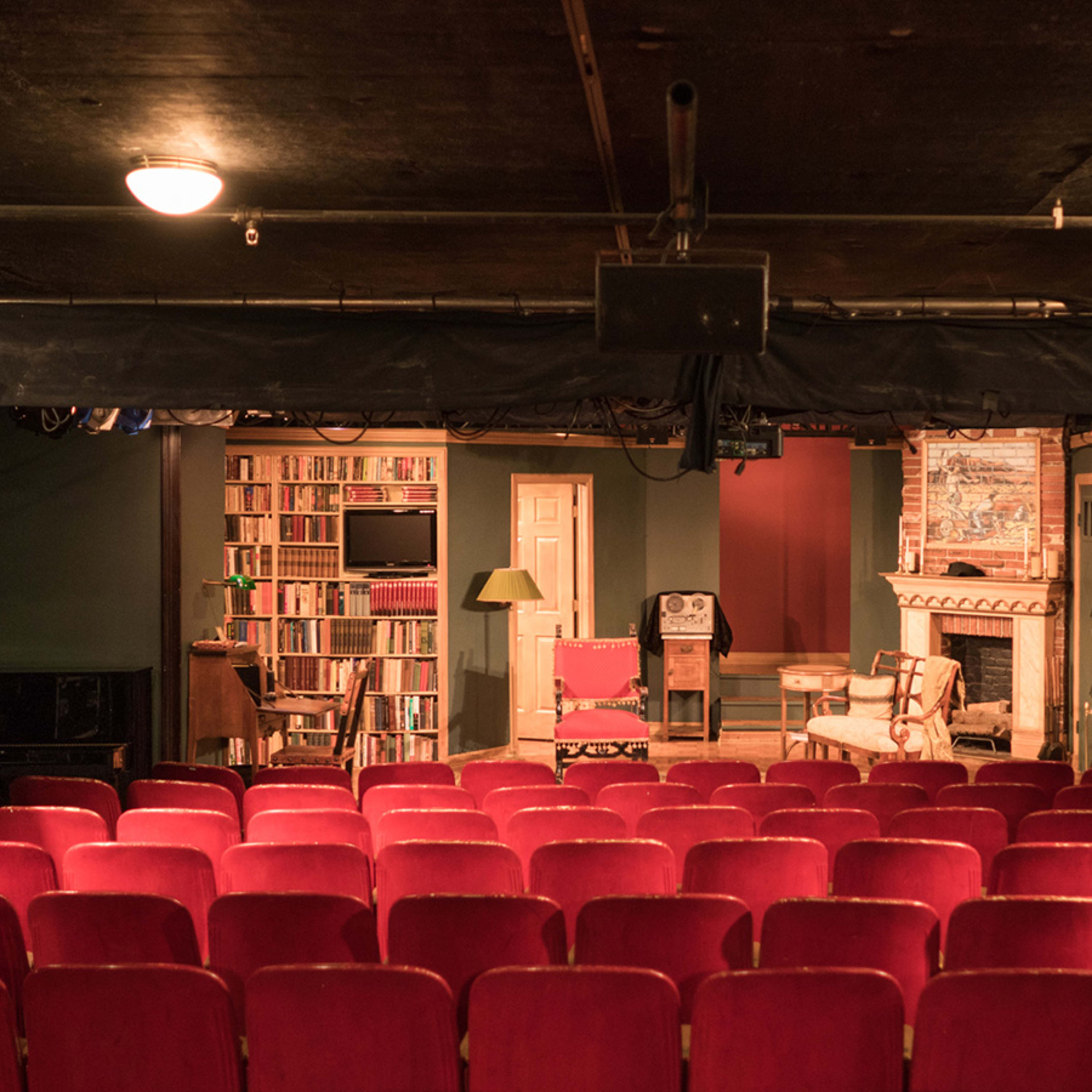 A small theater with red seating facing a stage that features a bookshelf, a fireplace, and a few chairs arranged for a performance.