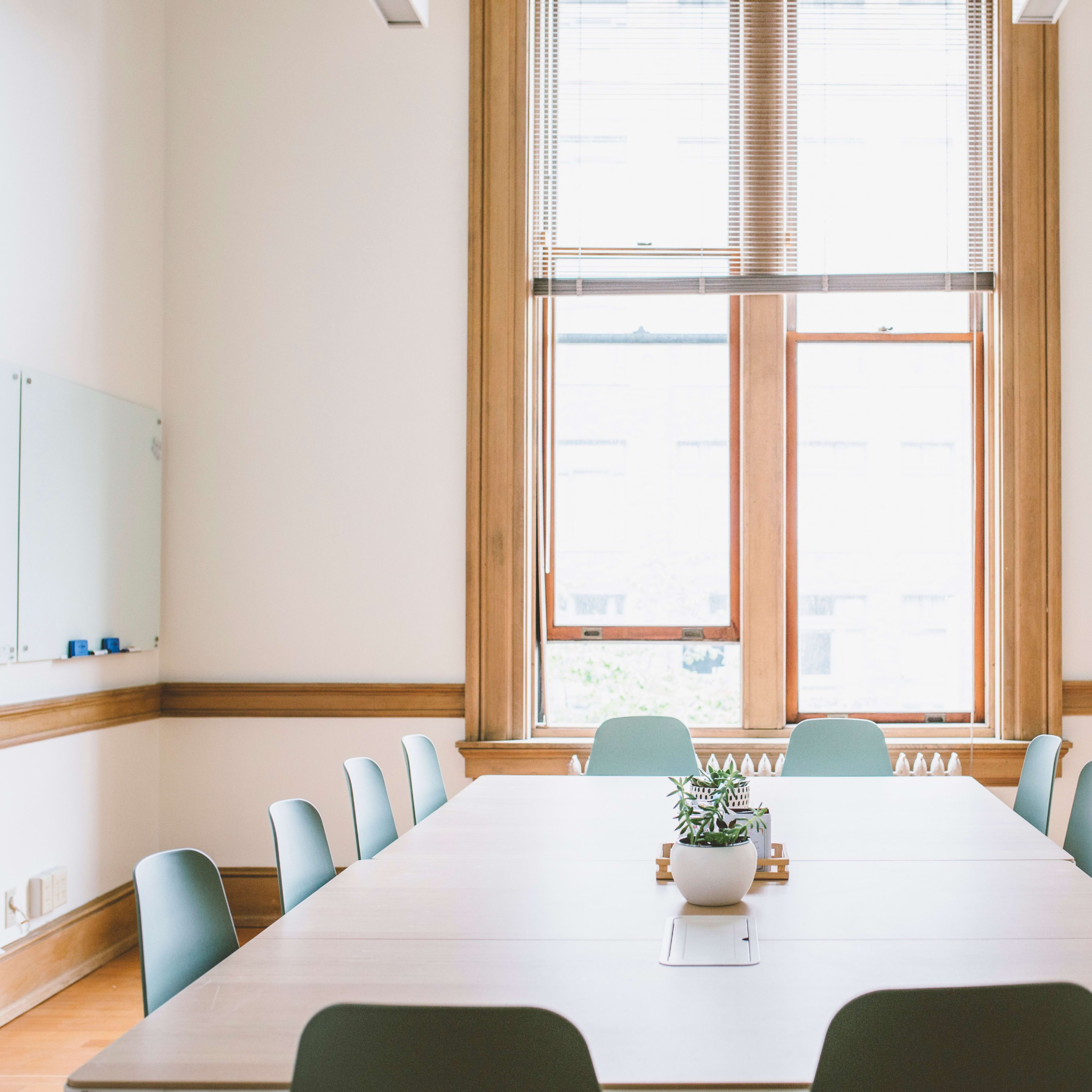A modern meeting room features a long wooden table surrounded by green chairs, with large windows letting in natural light.