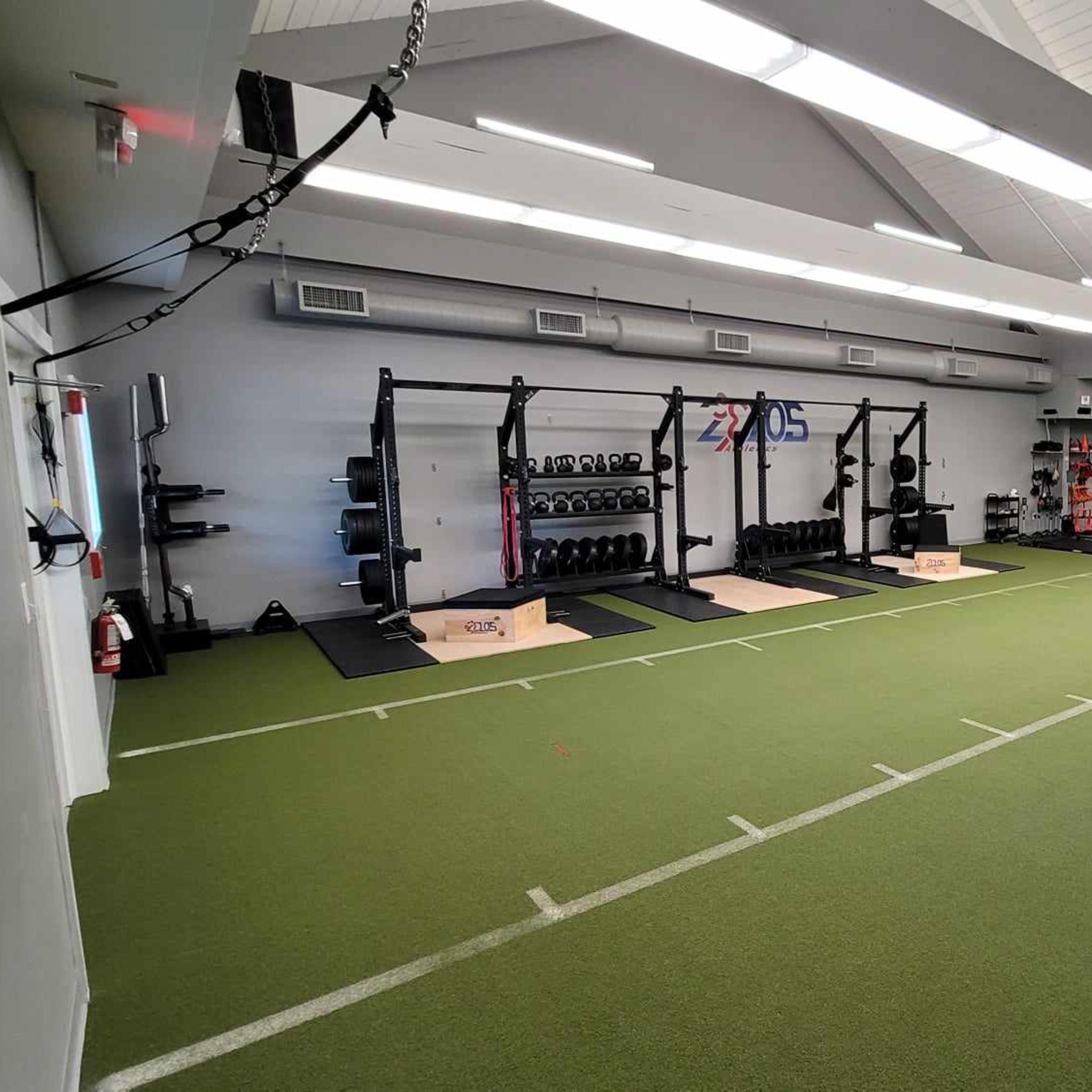 A spacious gym with turf flooring, multiple weightlifting stations, and exercise equipment arranged along the walls.