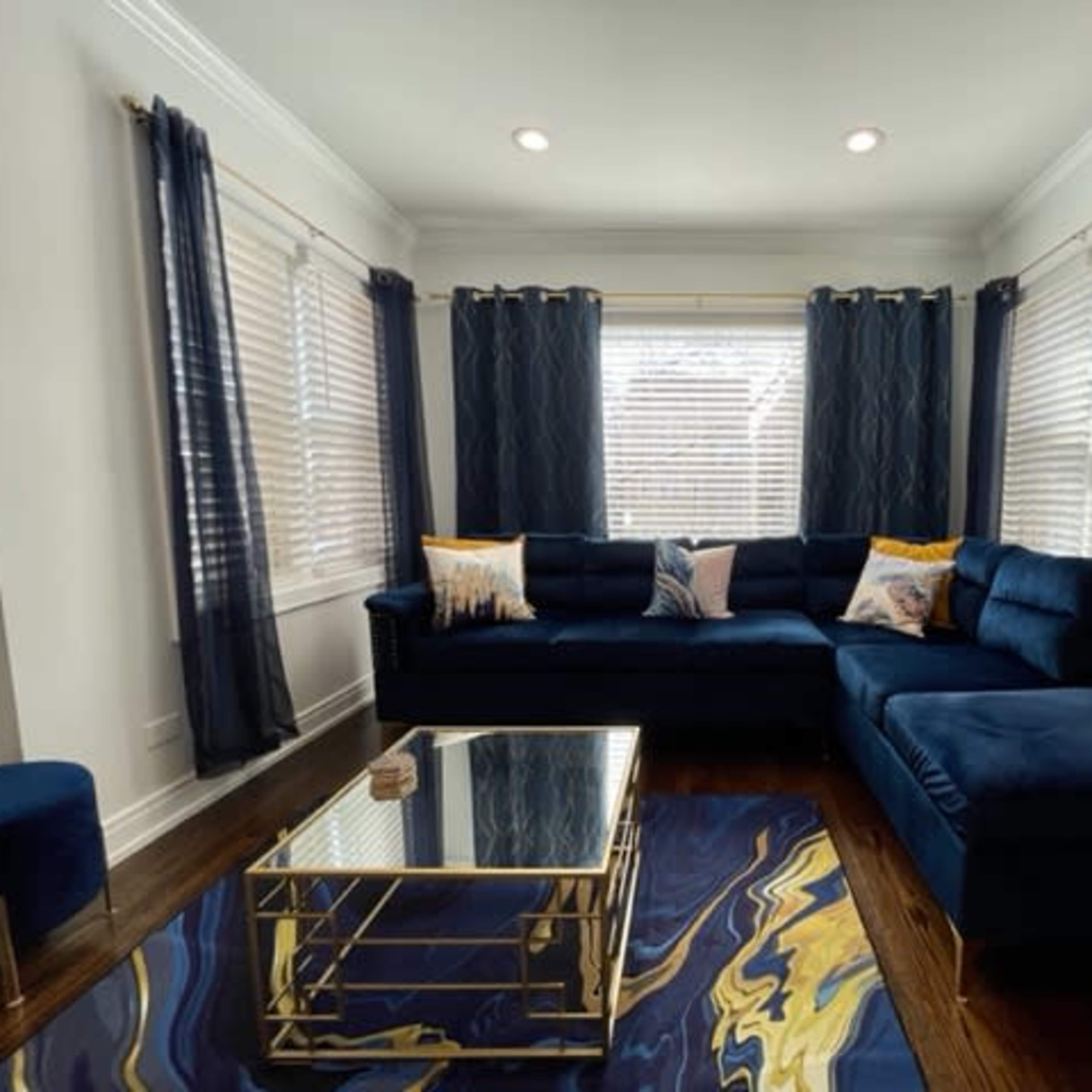 A living room with a blue sectional sofa, gold-accented coffee table, and large windows covered with dark curtains.
