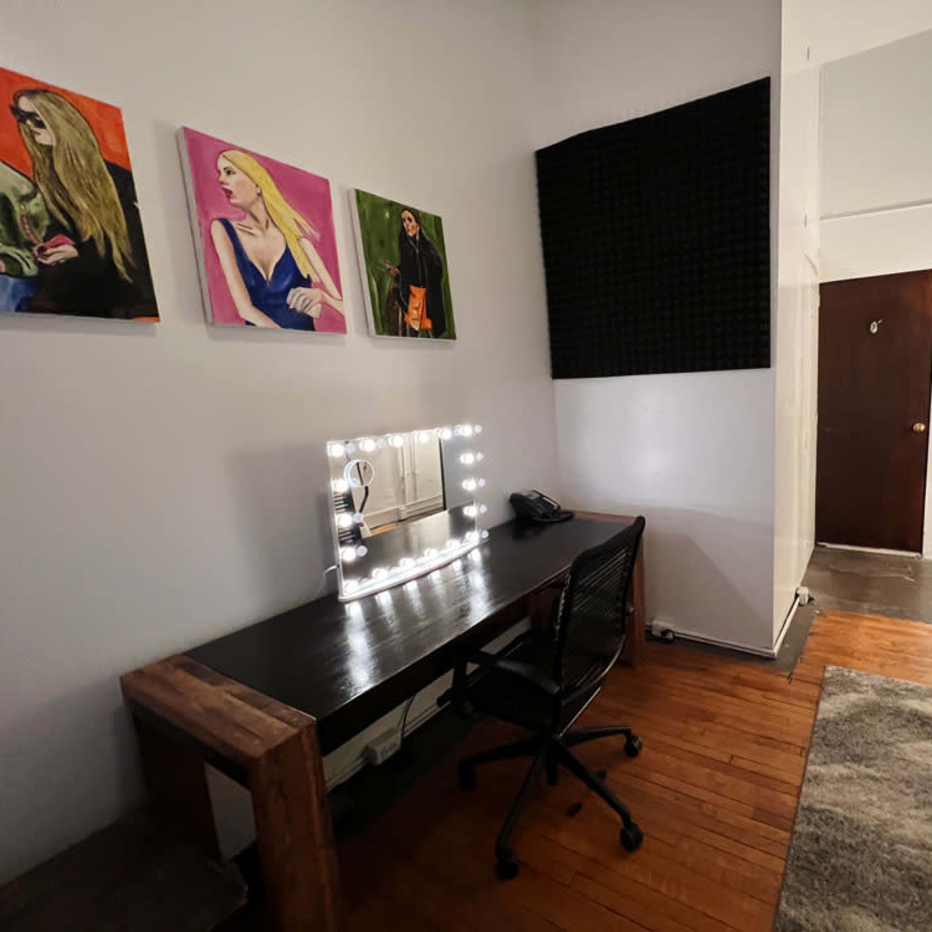 A small office or dressing area featuring a large illuminated mirror on a black desk, three colorful paintings on the wall, and a door leading to another room.