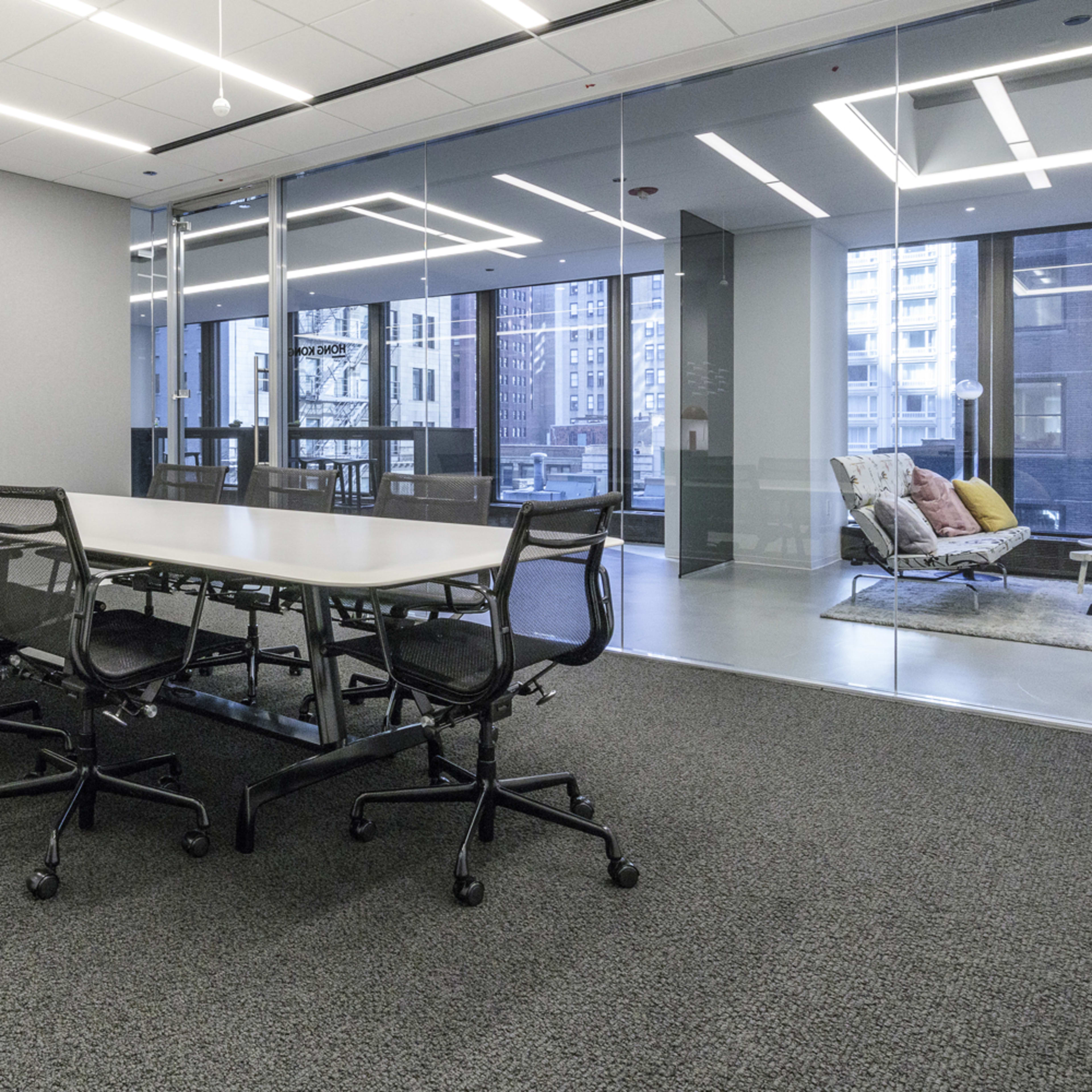 A modern conference room features a long table surrounded by ergonomic chairs, with a large screen mounted on one wall and glass windows providing a view of the city.