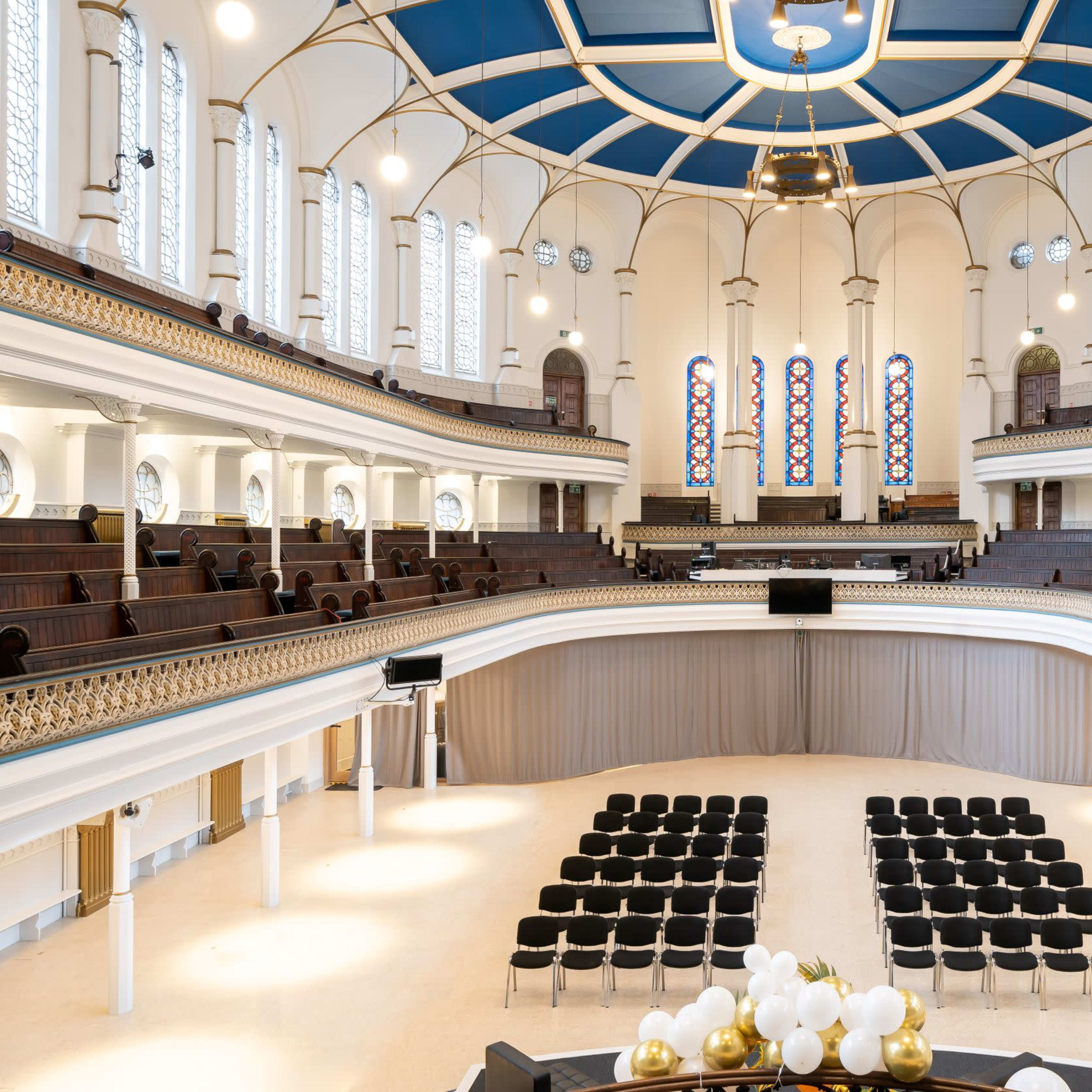 A spacious, elegantly designed auditorium with tiered seating arranged in front of a stage, featuring large windows and a decorative ceiling.