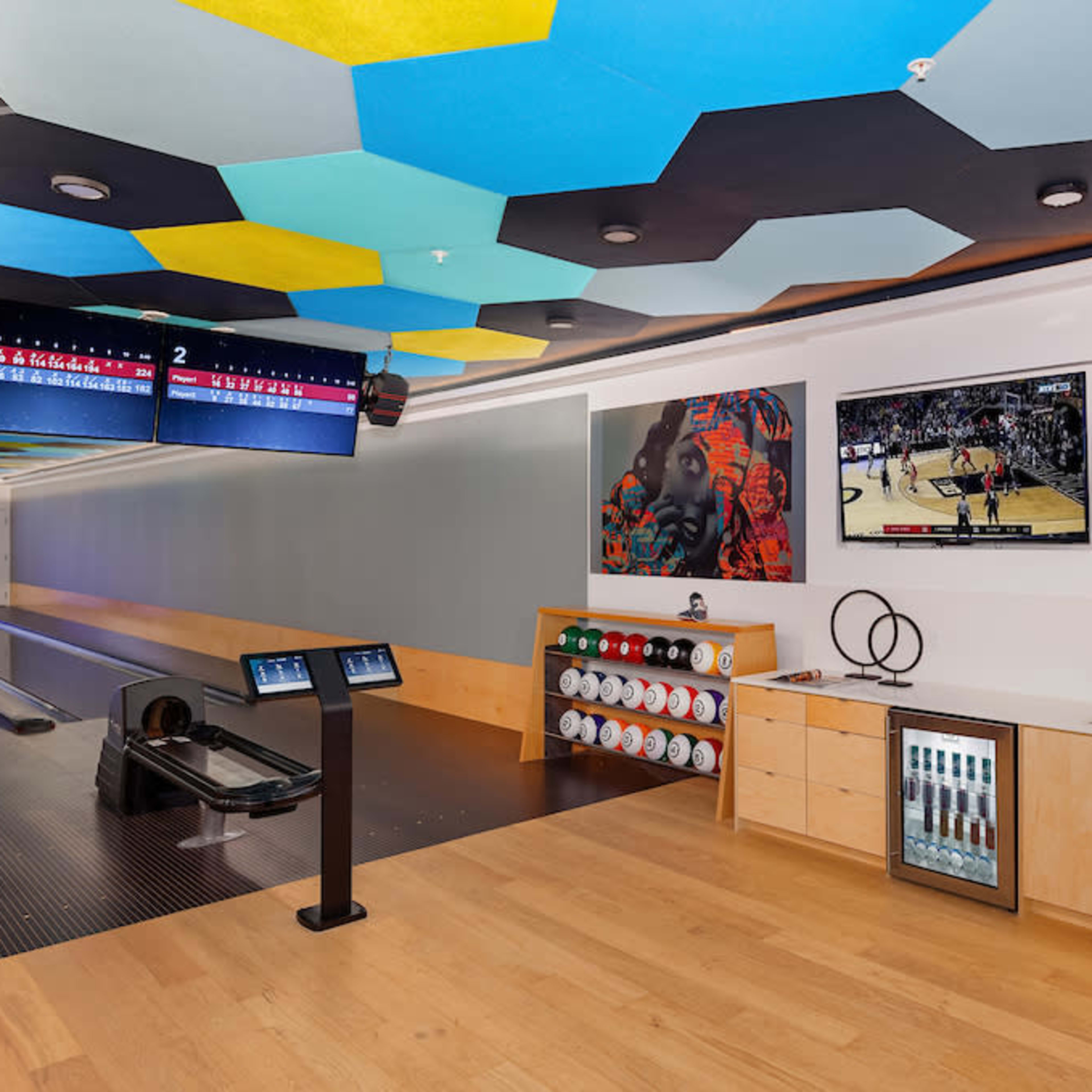 A modern bowling alley with two lanes, a seating area, two large televisions displaying a basketball game, and a colorful hexagonal ceiling.