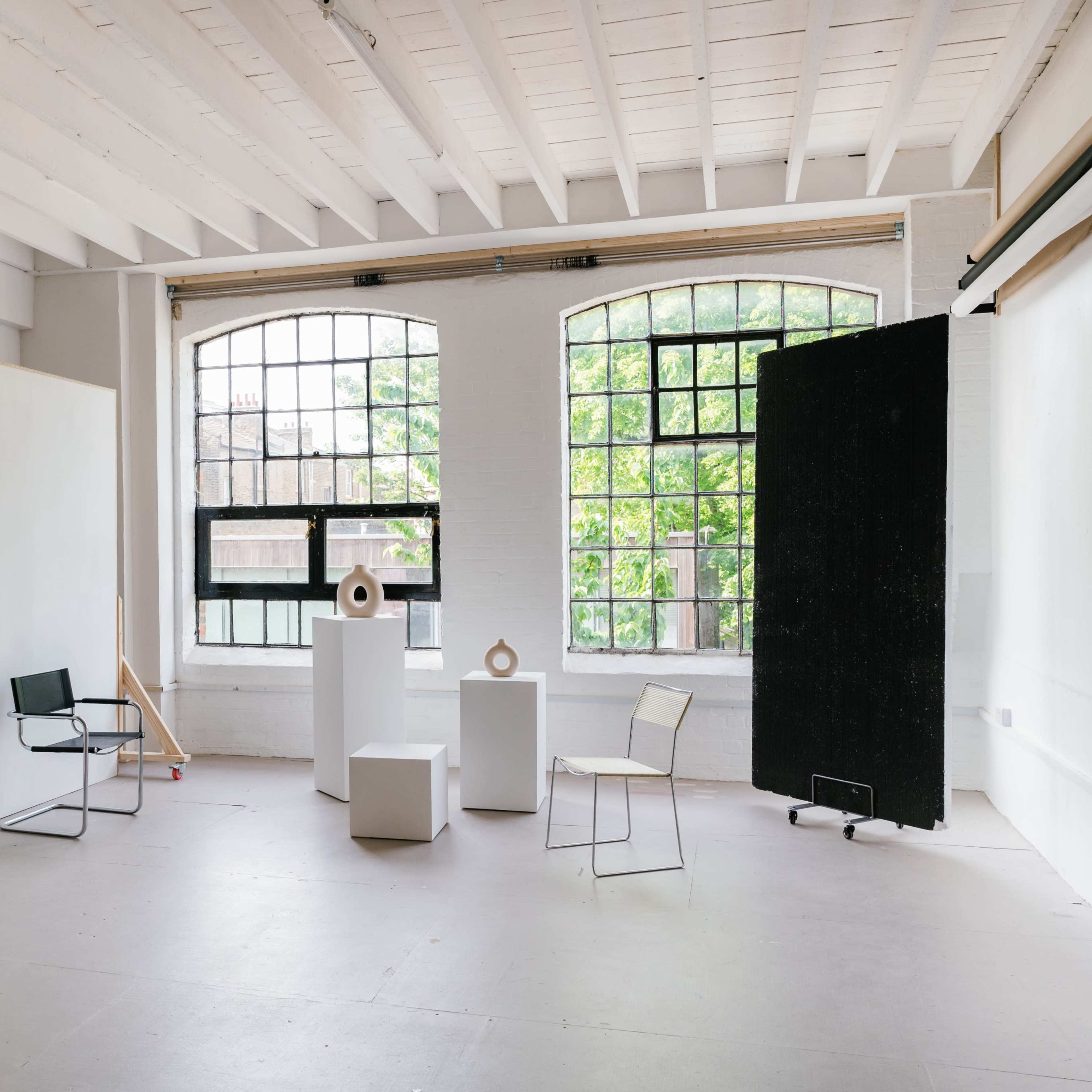 A spacious, well-lit studio featuring three white pedestals, two chairs, and large windows that overlook greenery.