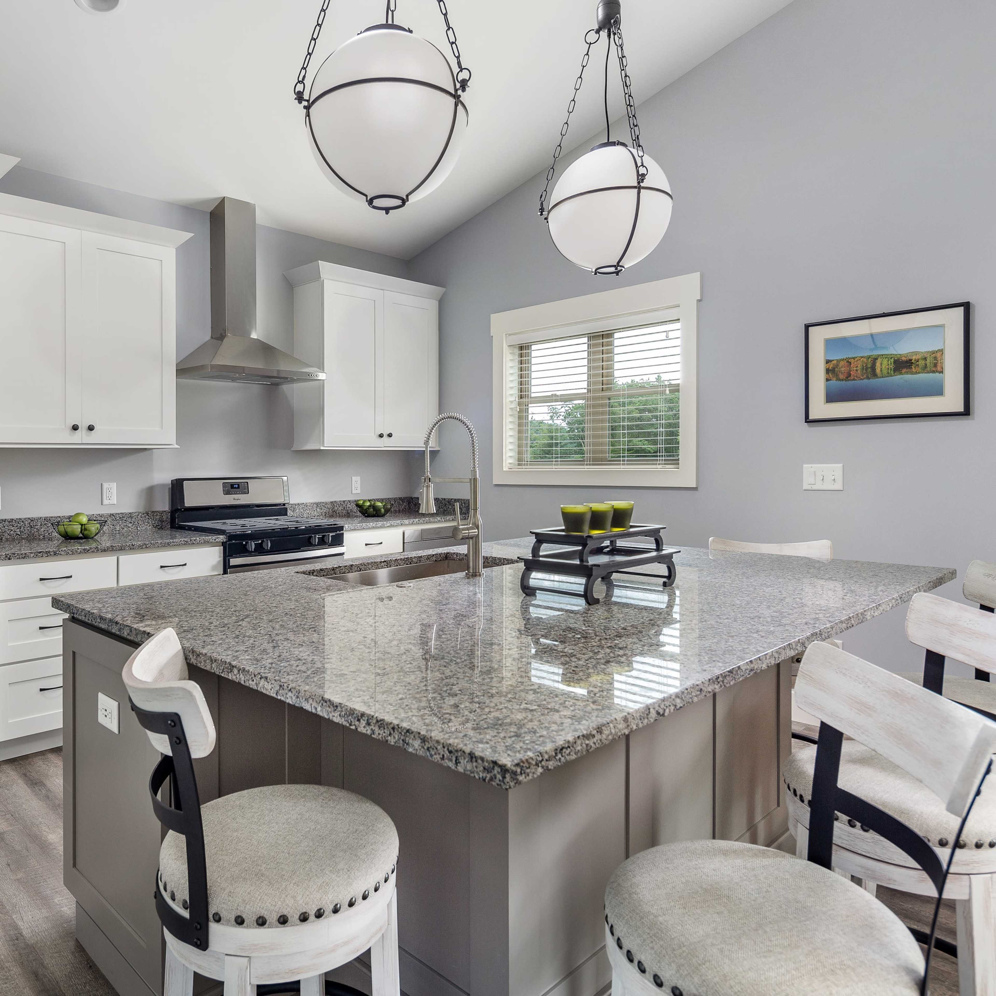 A modern kitchen features gray cabinets, a large granite island with seating, and stainless steel appliances.