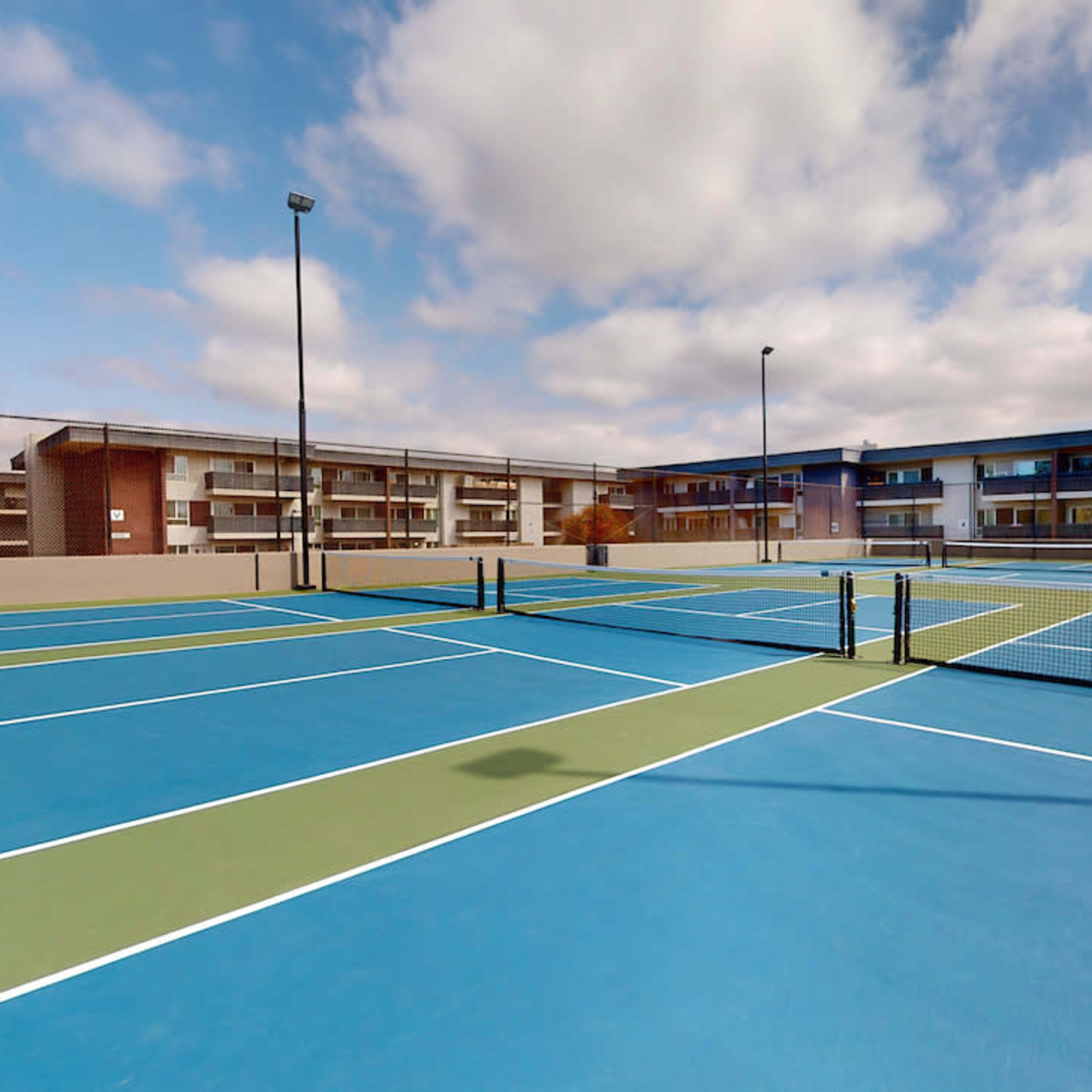 A series of blue and green tennis courts situated near a multi-story building under a partly cloudy sky.