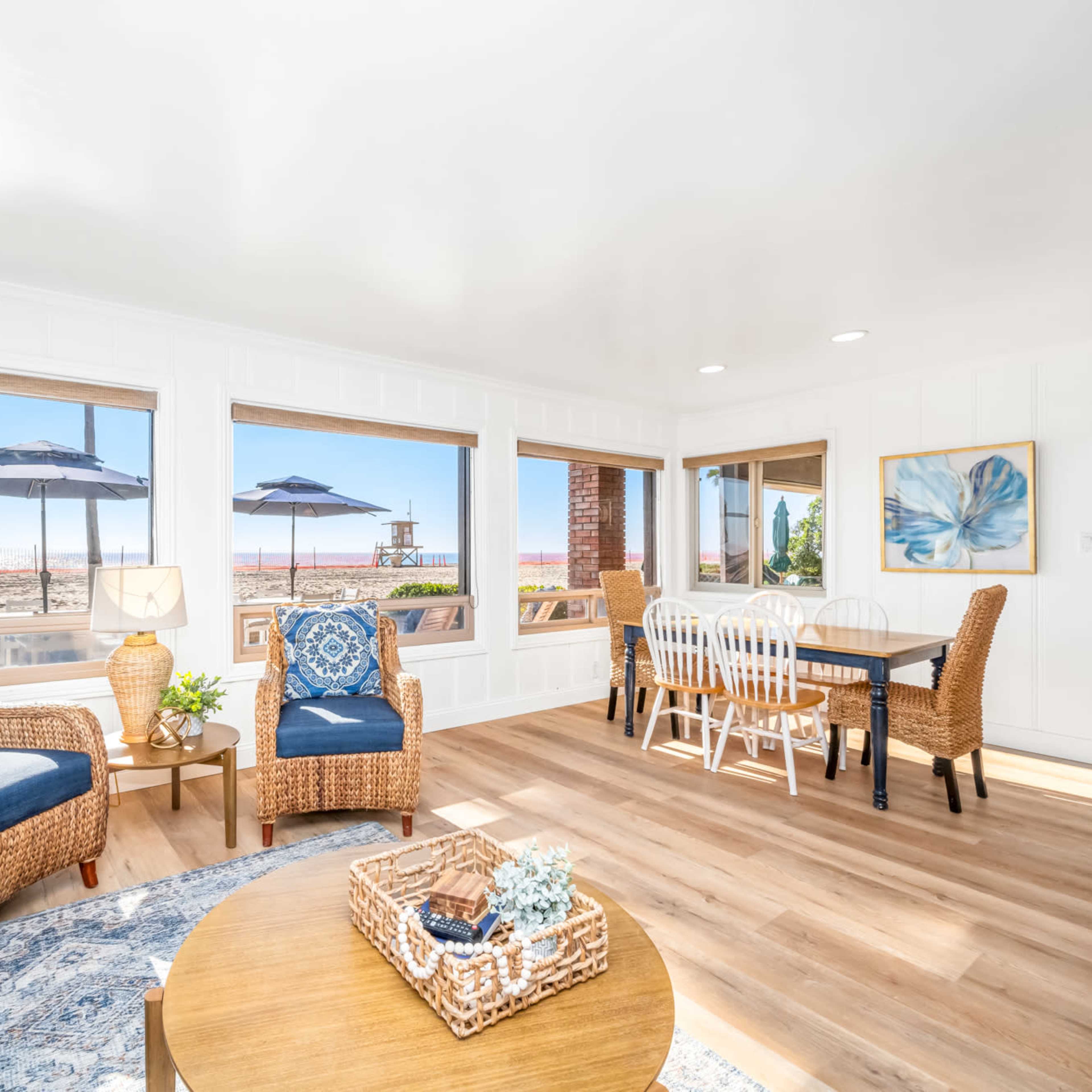 A bright living space features woven furniture, a round wooden table, and large windows showcasing a beach view with umbrellas outside.