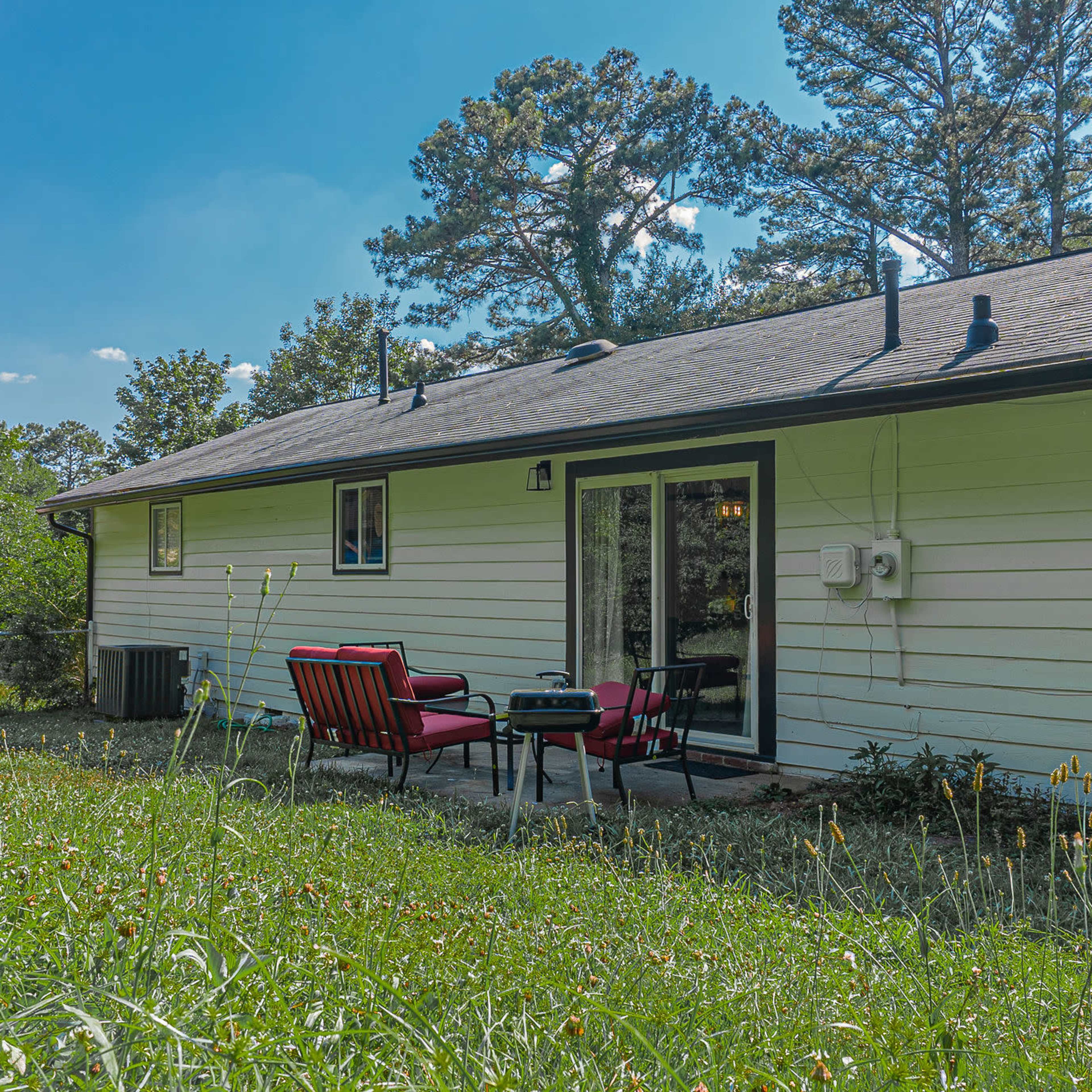 A backyard scene featuring a single-story house with a patio area, surrounded by grass and trees.