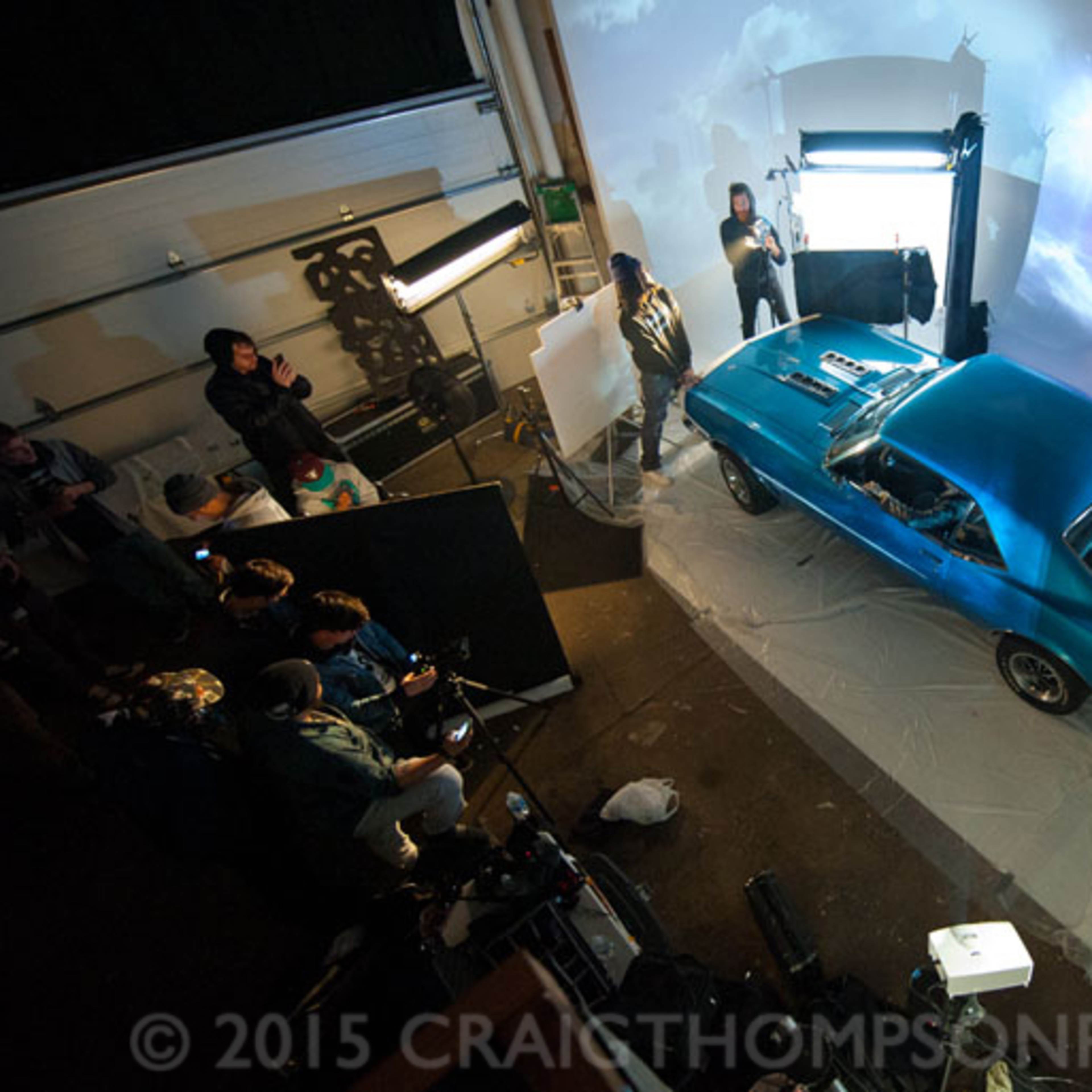 A blue vintage car is being photographed in a studio setting with multiple lights and crew members arranging equipment around it.