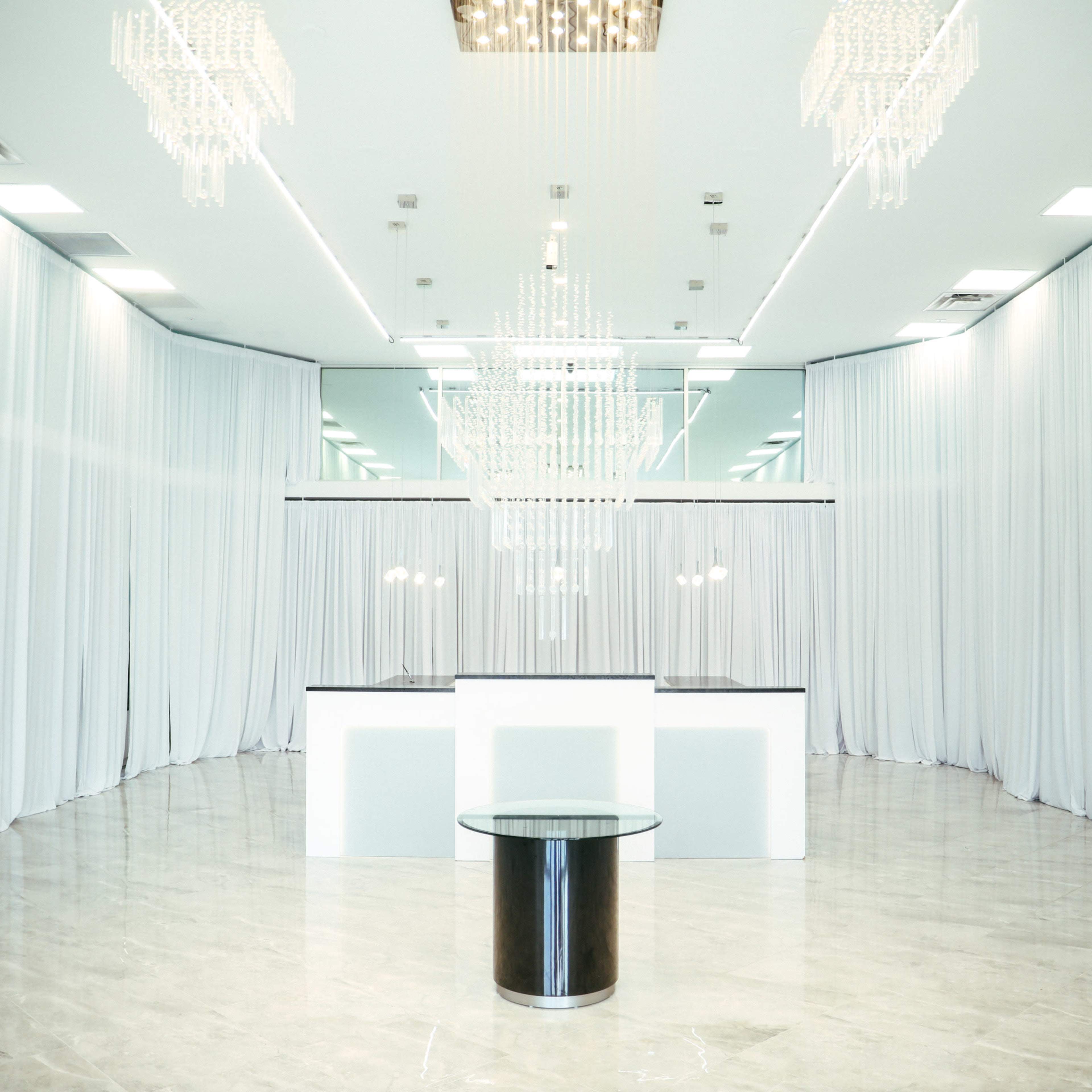 An empty event space featuring white curtain drapery, two large chandeliers, and a central round table.