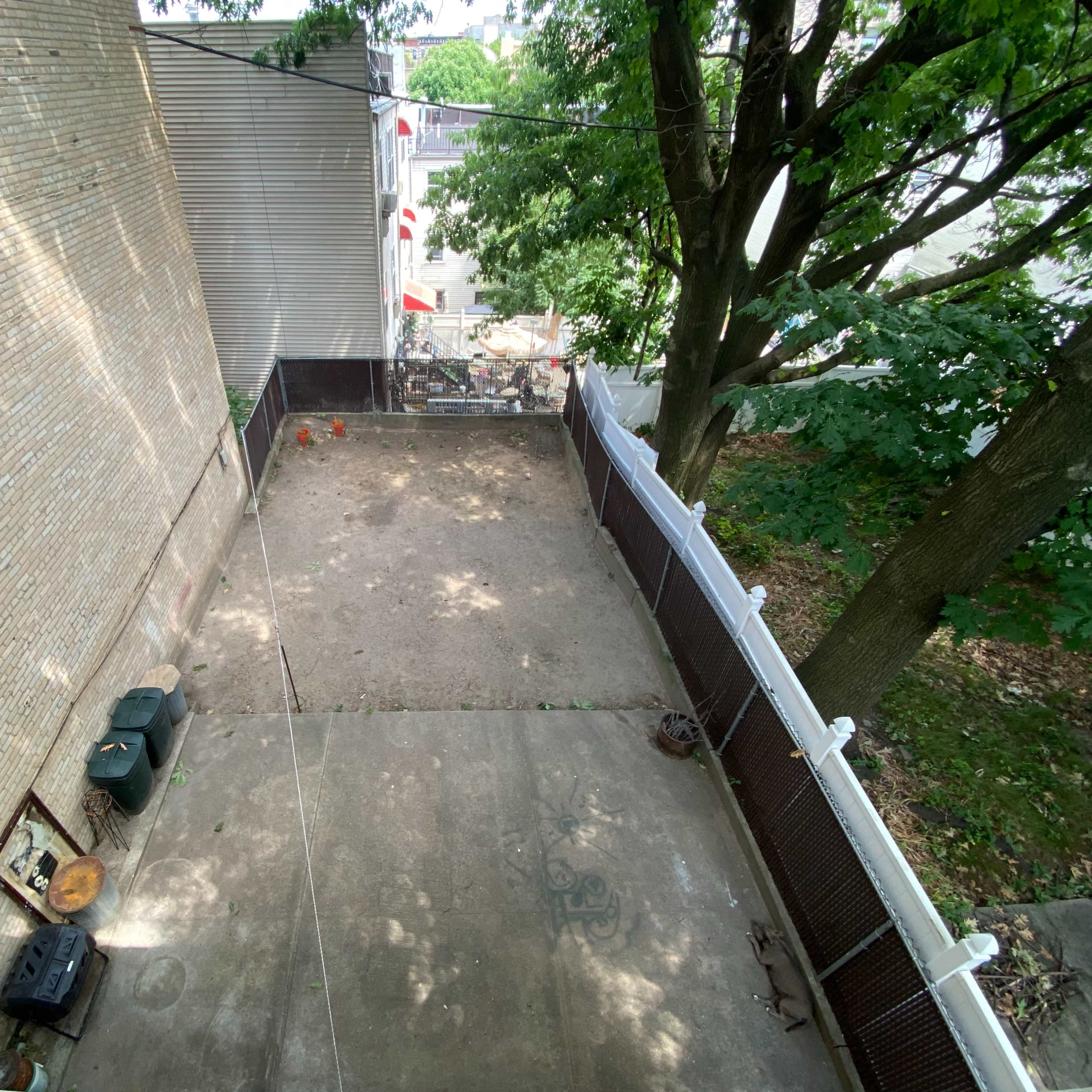 An empty backyard area with concrete flooring, surrounded by a fence and a tree on one side.