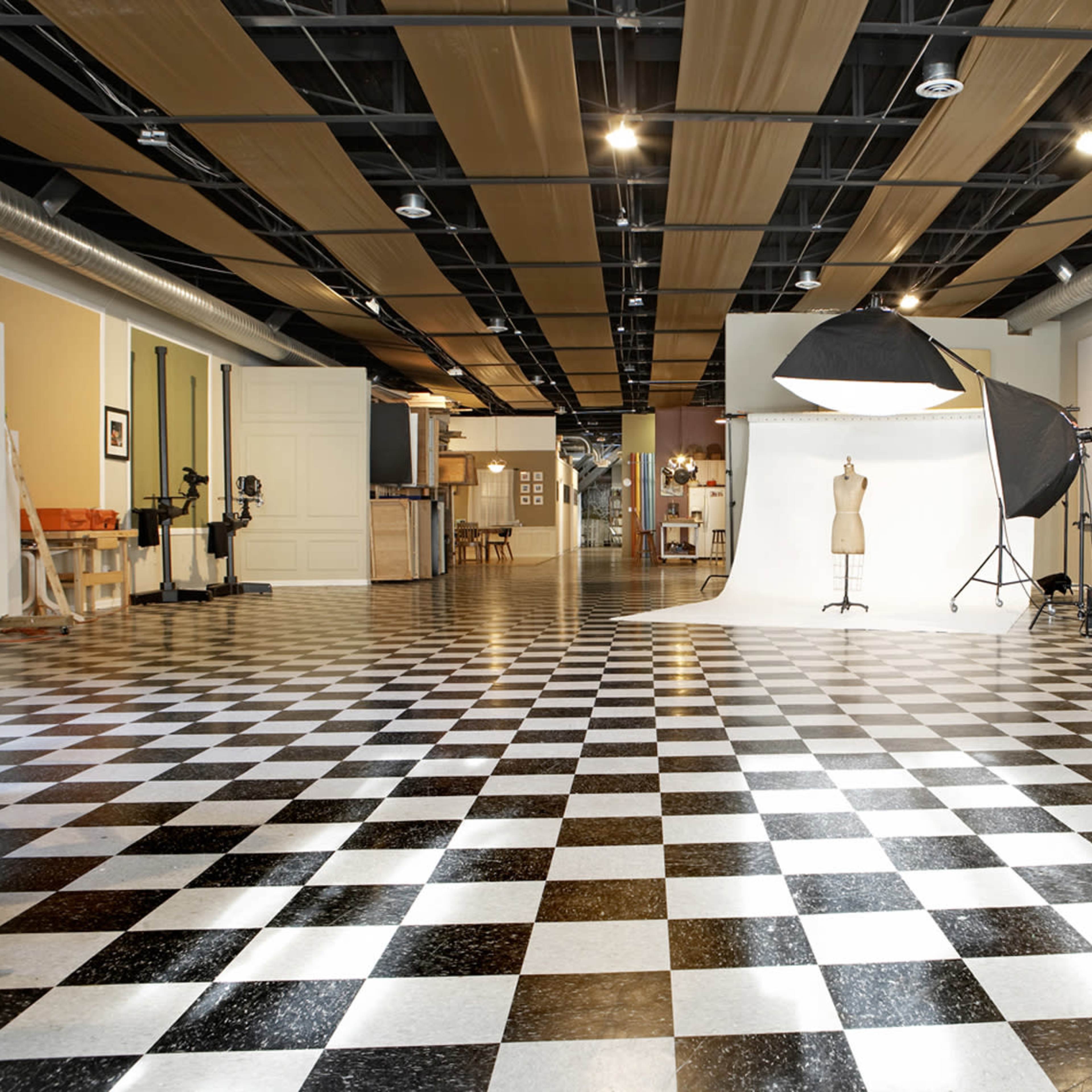 A spacious photography studio with a checkered floor, two white backdrop stands, a mannequin, and various equipment set up.