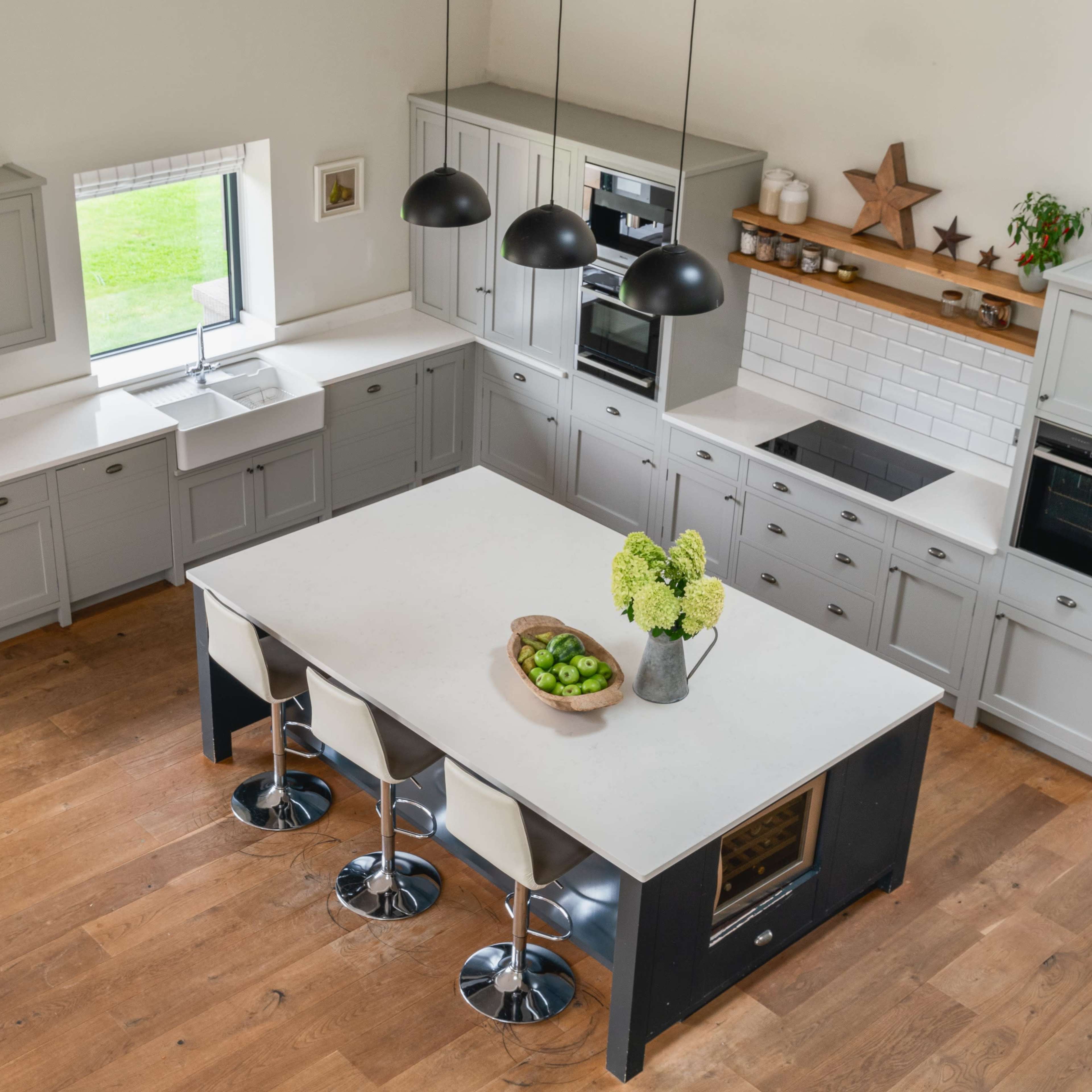 A modern kitchen with gray cabinetry, a large central island, and sleek bar stools, featuring wooden flooring and large windows.