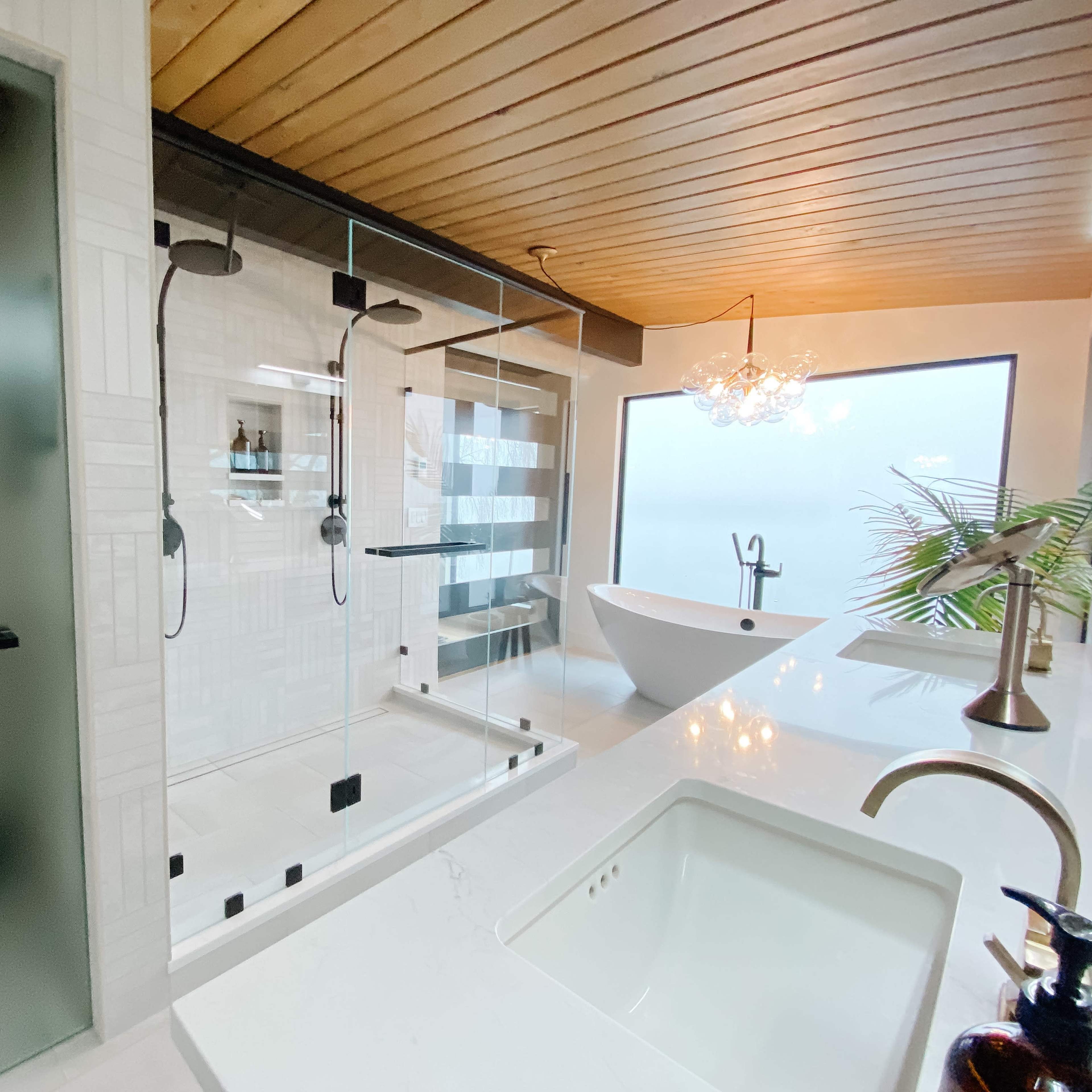 A modern bathroom featuring a glass shower enclosure, a freestanding bathtub, a double sink vanity, and large windows providing natural light.