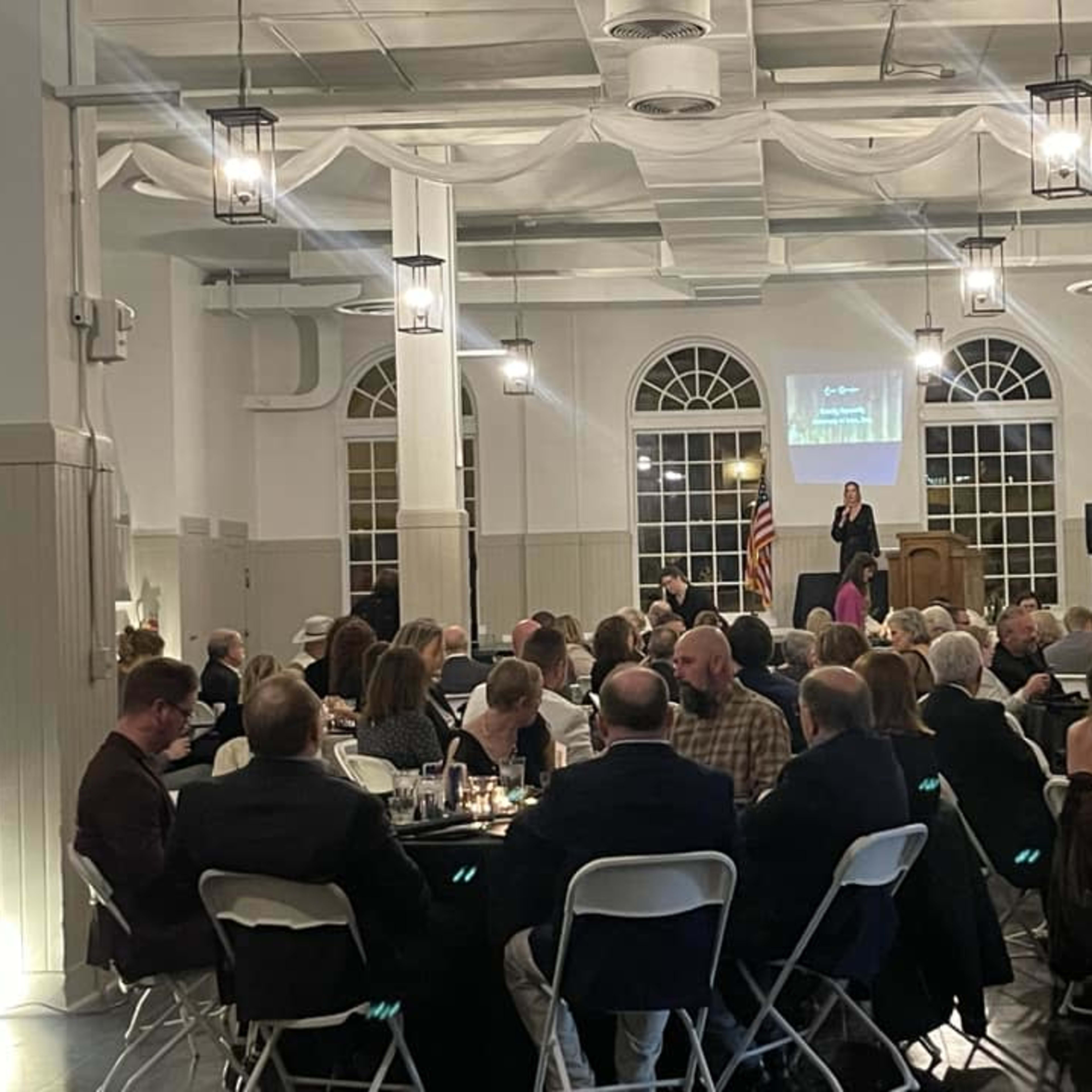 A group of people attend a formal event in a large, well-lit hall with tables set for dining and a speaker at a podium in the background.
