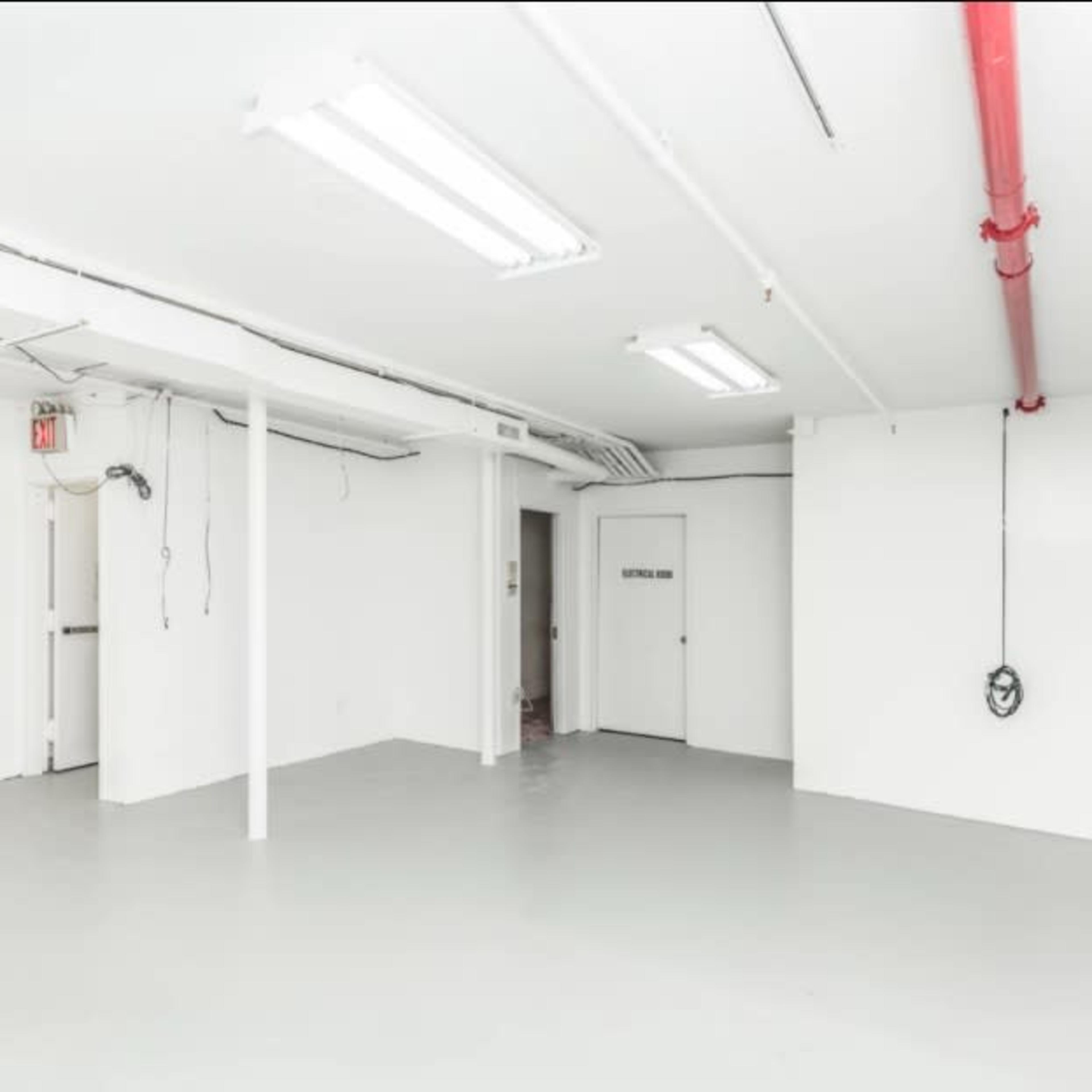 A spacious, empty room with white walls and a gray floor, featuring overhead fluorescent lights and a red pipe along the ceiling.