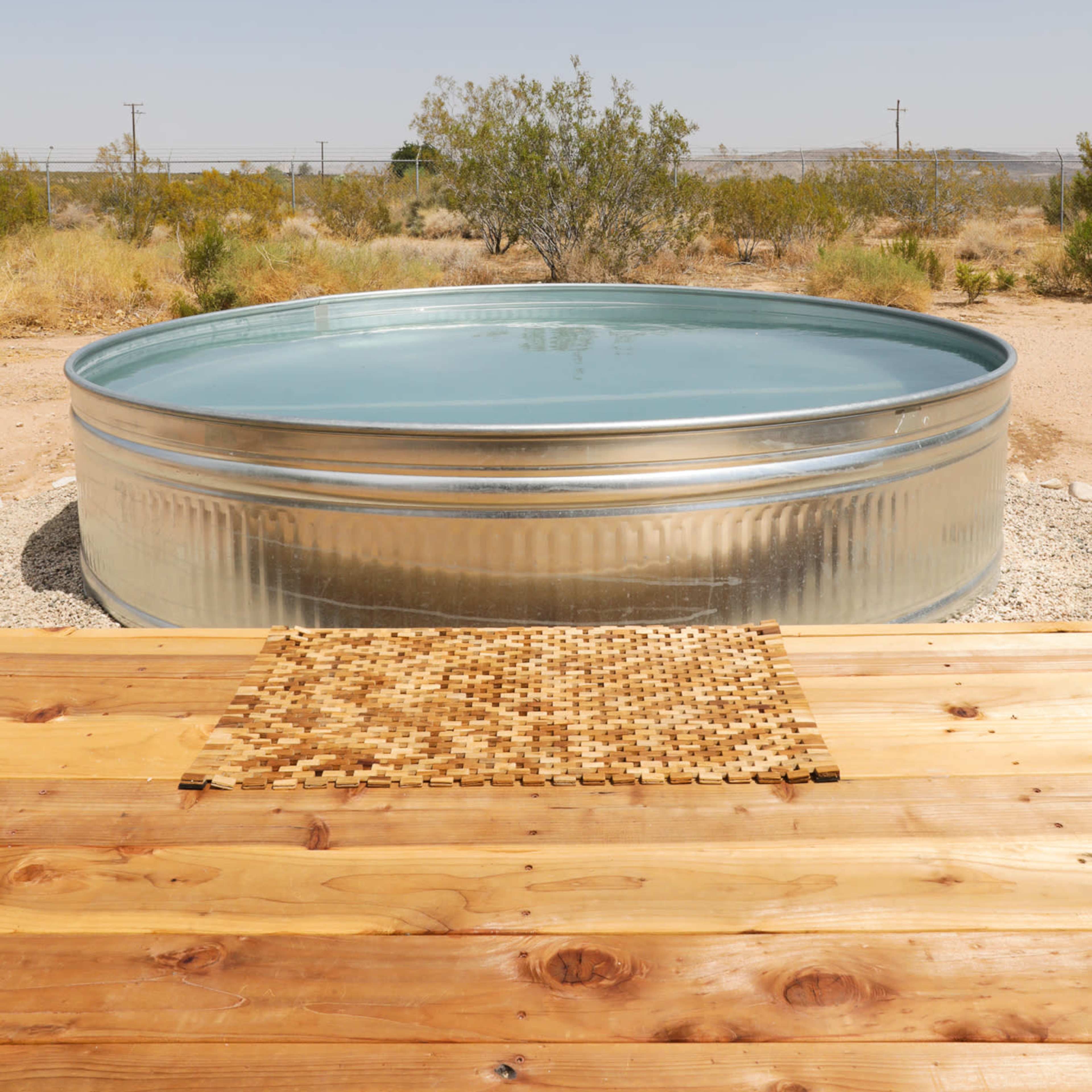 A large, circular metal water tank sits filled with water on a gravel surface, surrounded by sparse desert vegetation under a clear blue sky.