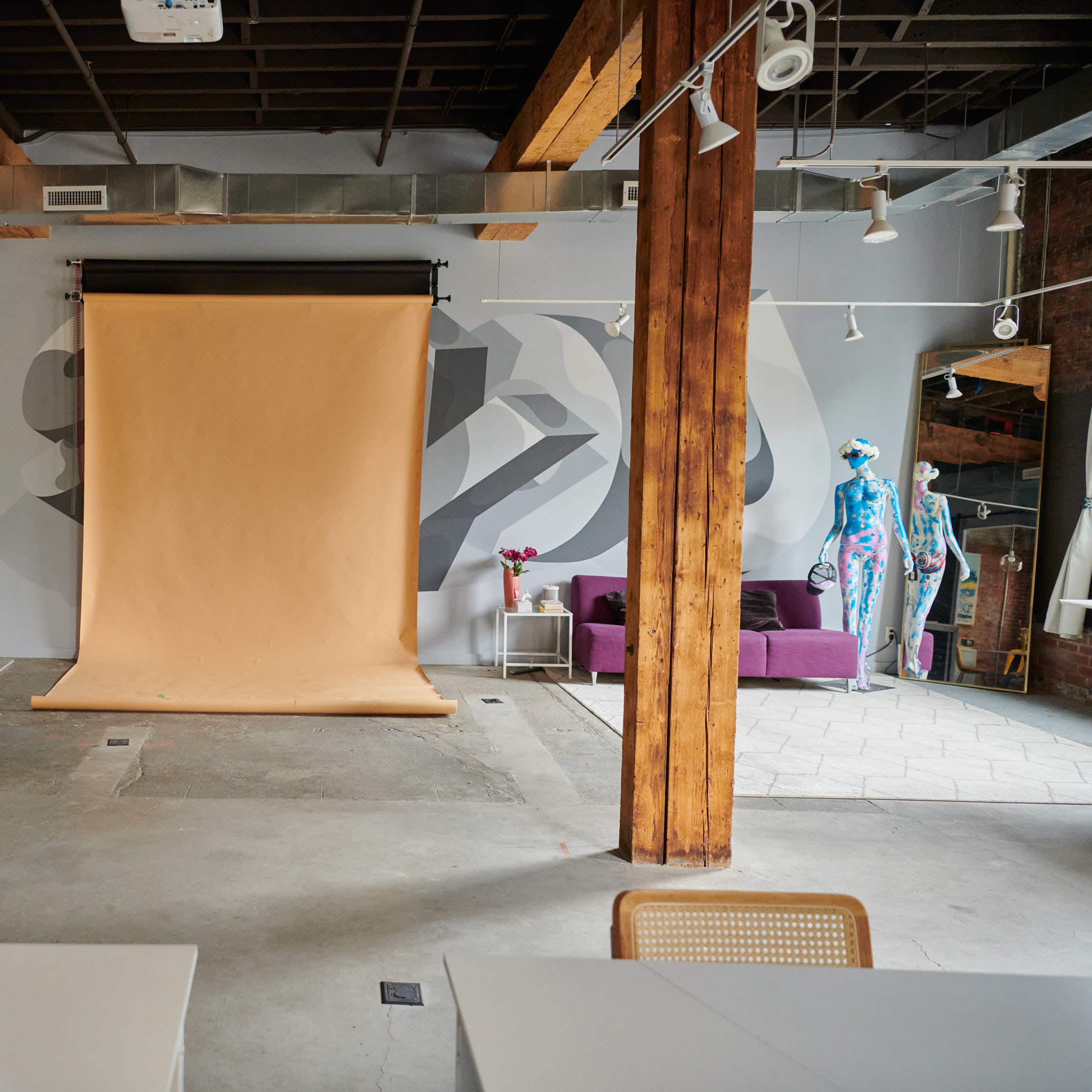 An industrial-style studio with wooden columns, a tan backdrop, and a purple couch positioned against a mural on the wall, along with tables and chairs arranged in the space.