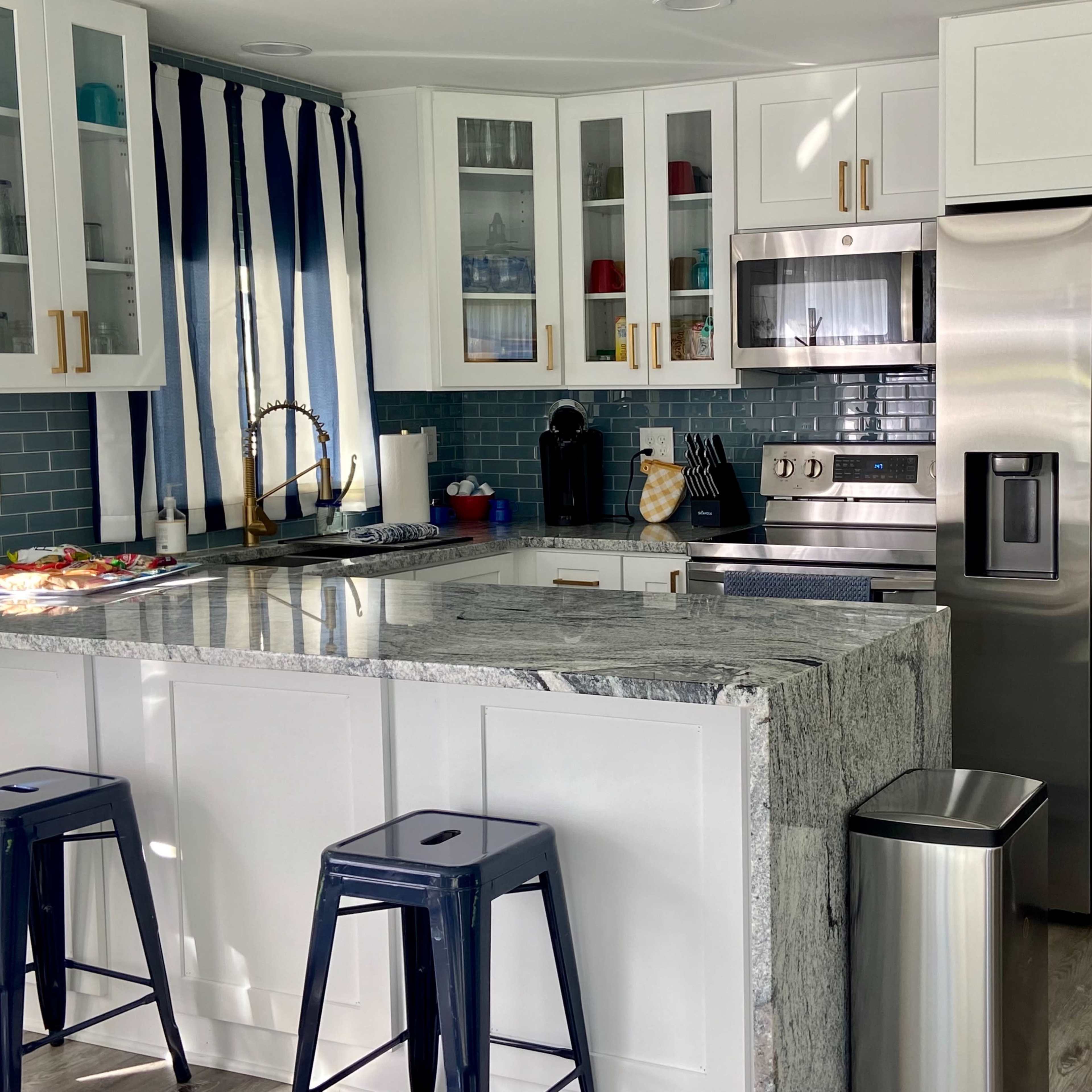 A modern kitchen with a white island, stainless steel appliances, blue and white cabinetry, and a bar area with two metal stools.
