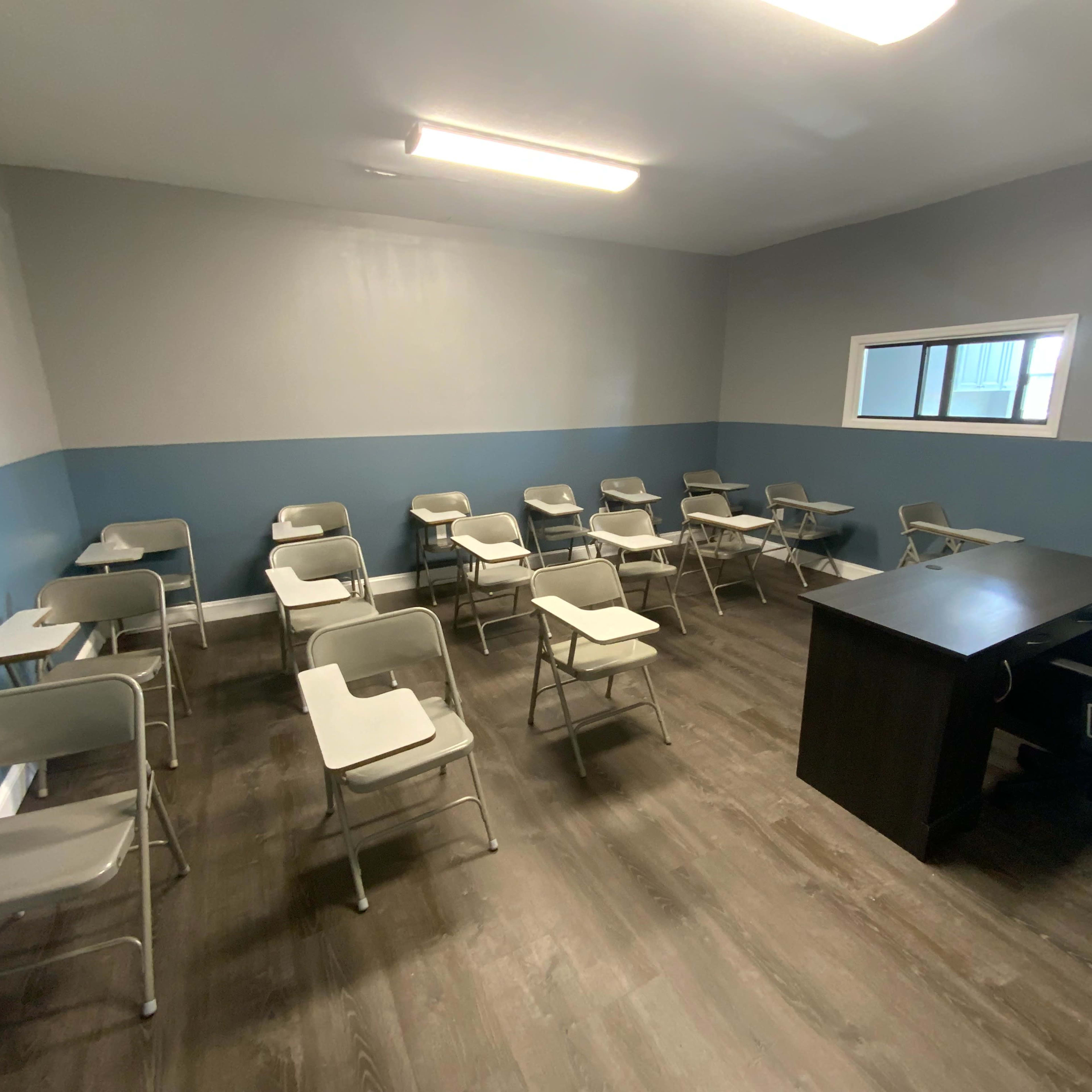 A sparse classroom arranged with individual desks facing a single teacher's desk against a gray and blue wall.