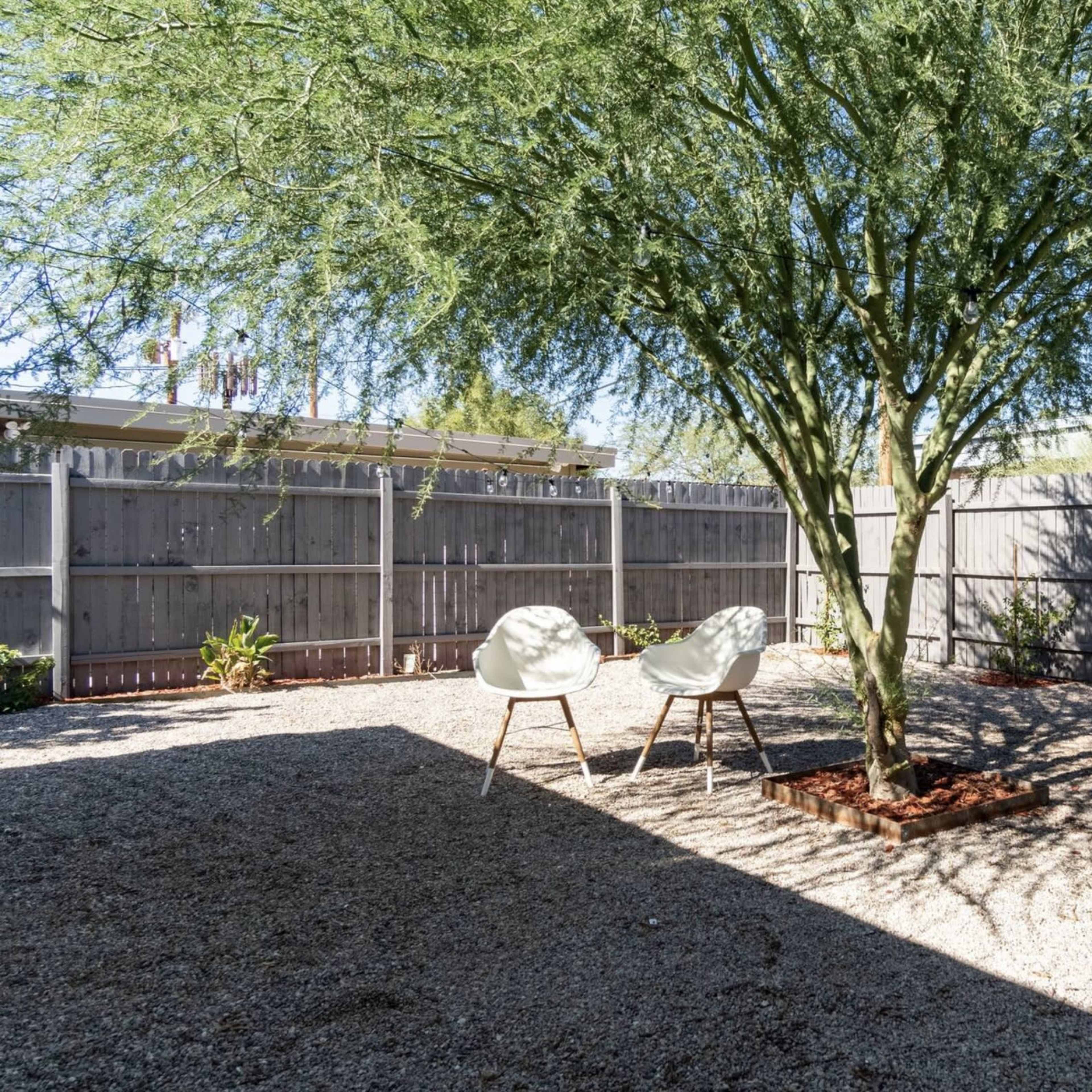 A shaded outdoor space features two white chairs set on gravel beneath a tree, surrounded by a wooden fence.