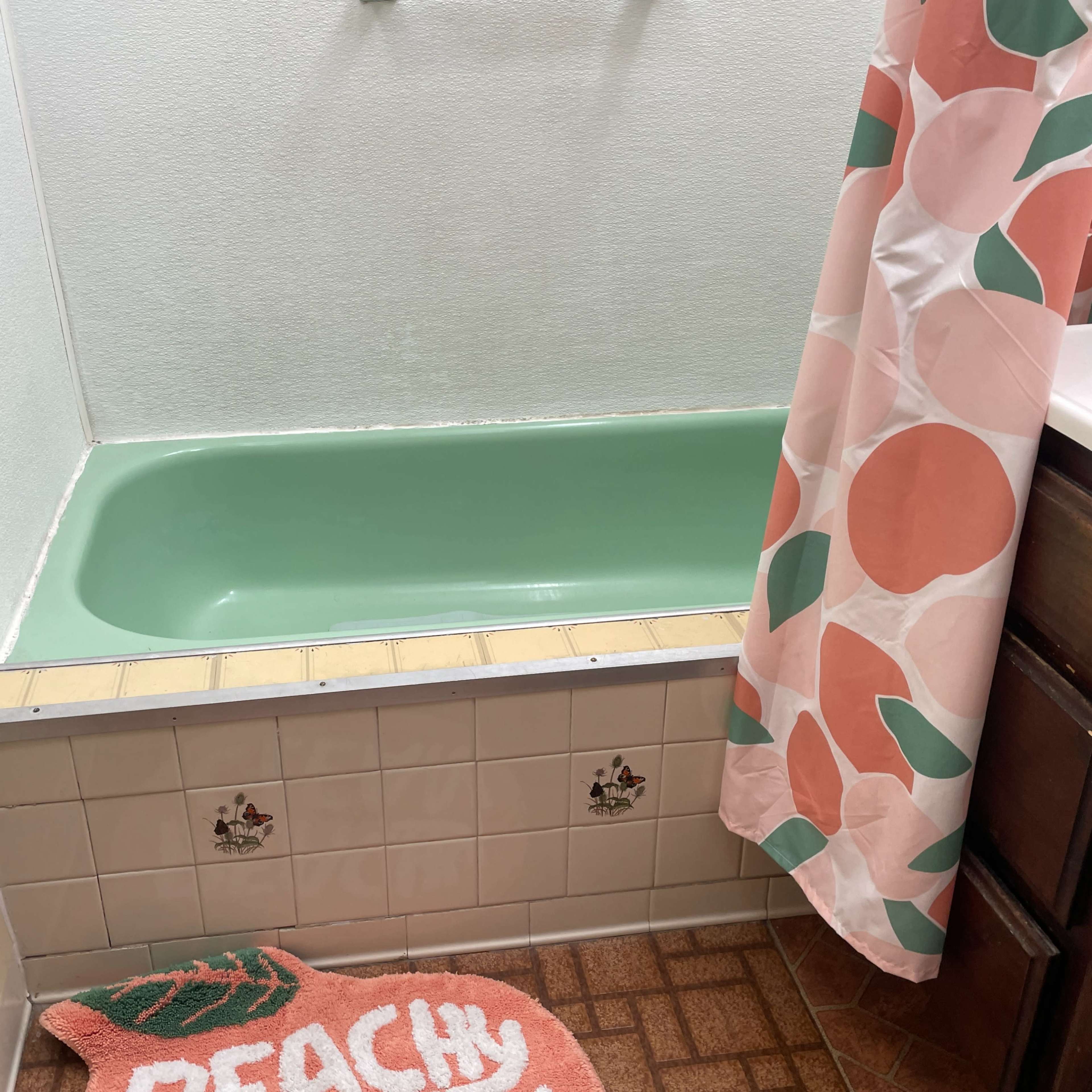 A bathroom featuring a mint green bathtub, a patterned shower curtain, and a peach-themed bath mat on a tiled floor.