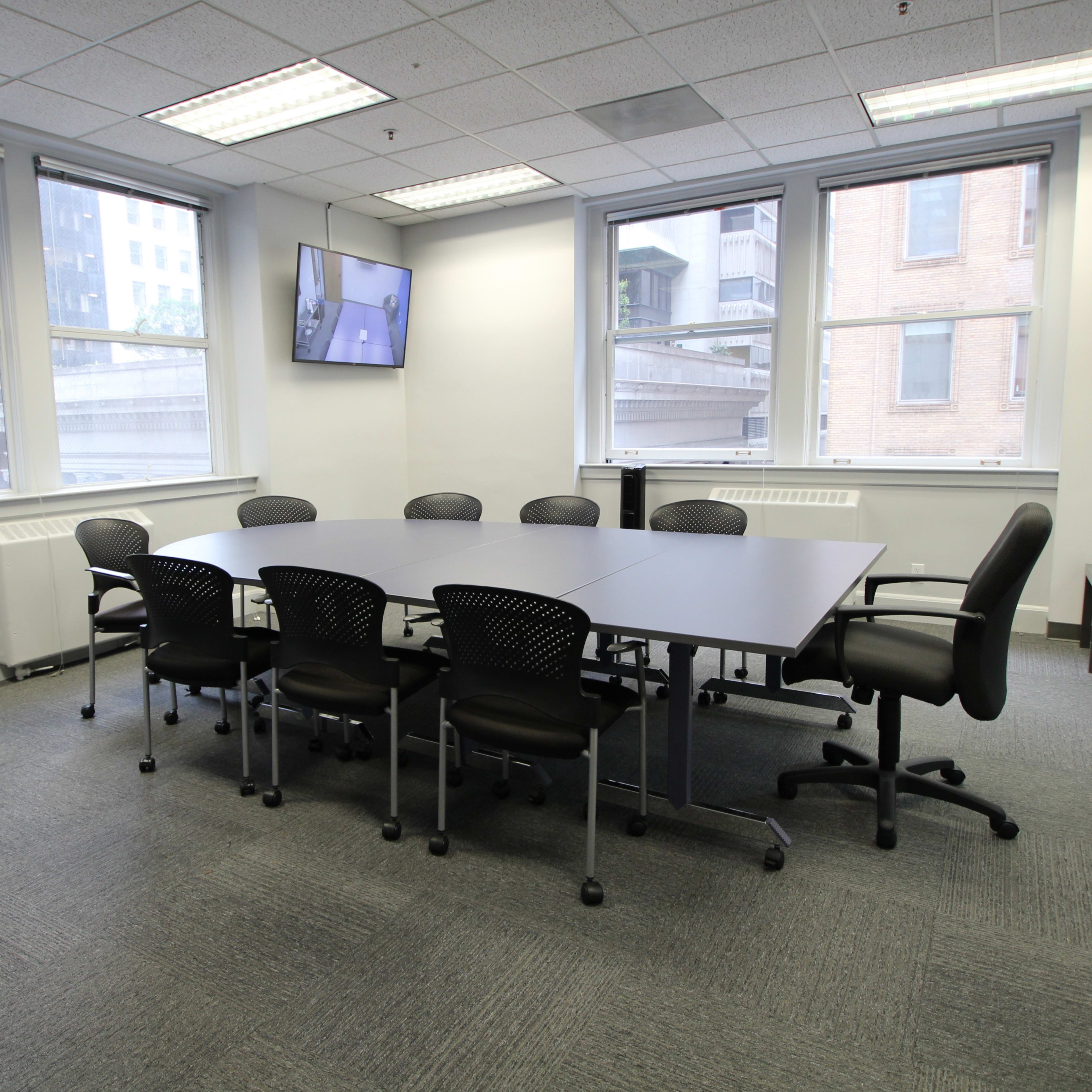 A conference room with a large table surrounded by chairs, featuring a wall-mounted television and large windows.