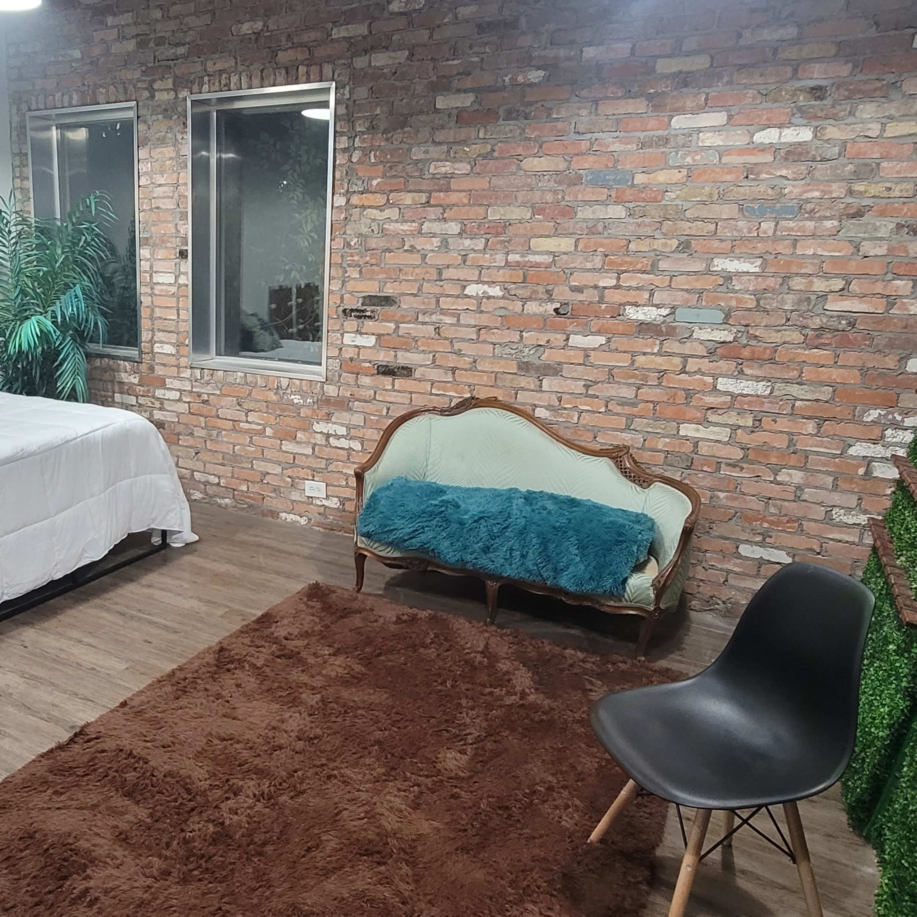 The room features a bed with white bedding, a green couch, a brown rug, and a brick wall backdrop with plants arranged on one side.
