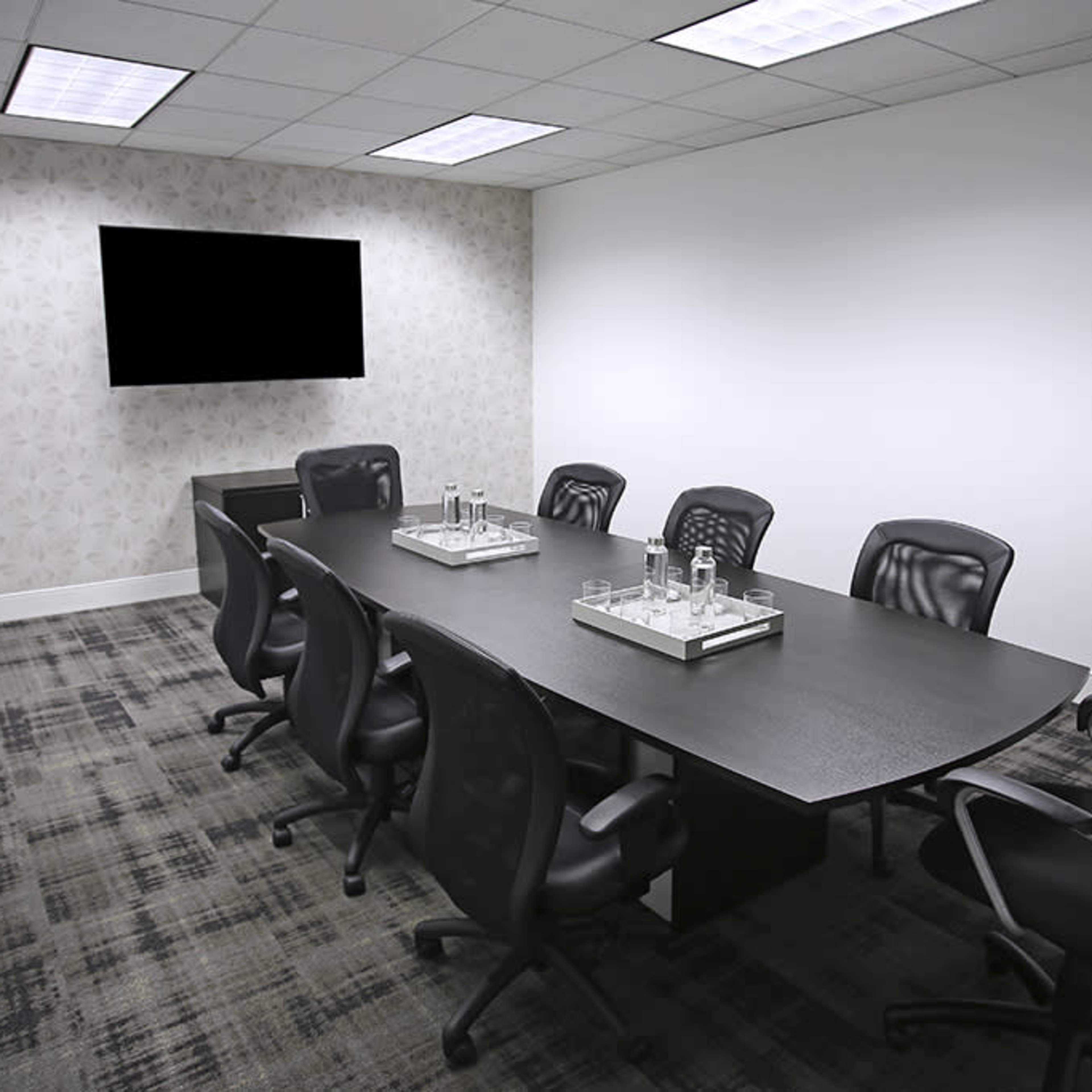 A modern conference room with a large table surrounded by ergonomic chairs, a television mounted on the wall, and a patterned backdrop.