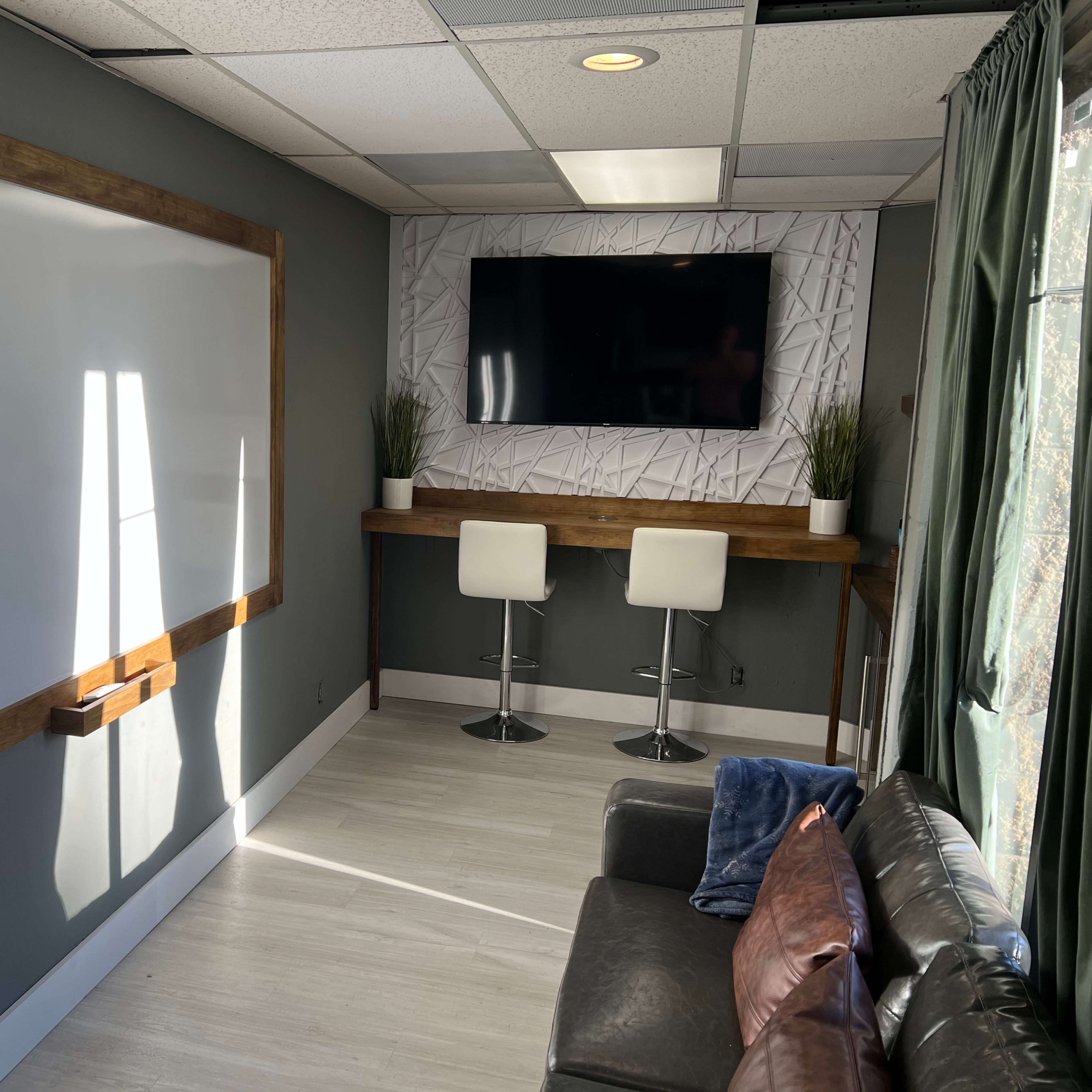 A small meeting room with a whiteboard, a TV mounted on the wall, a wooden counter with two white stools, and a couch in the corner.