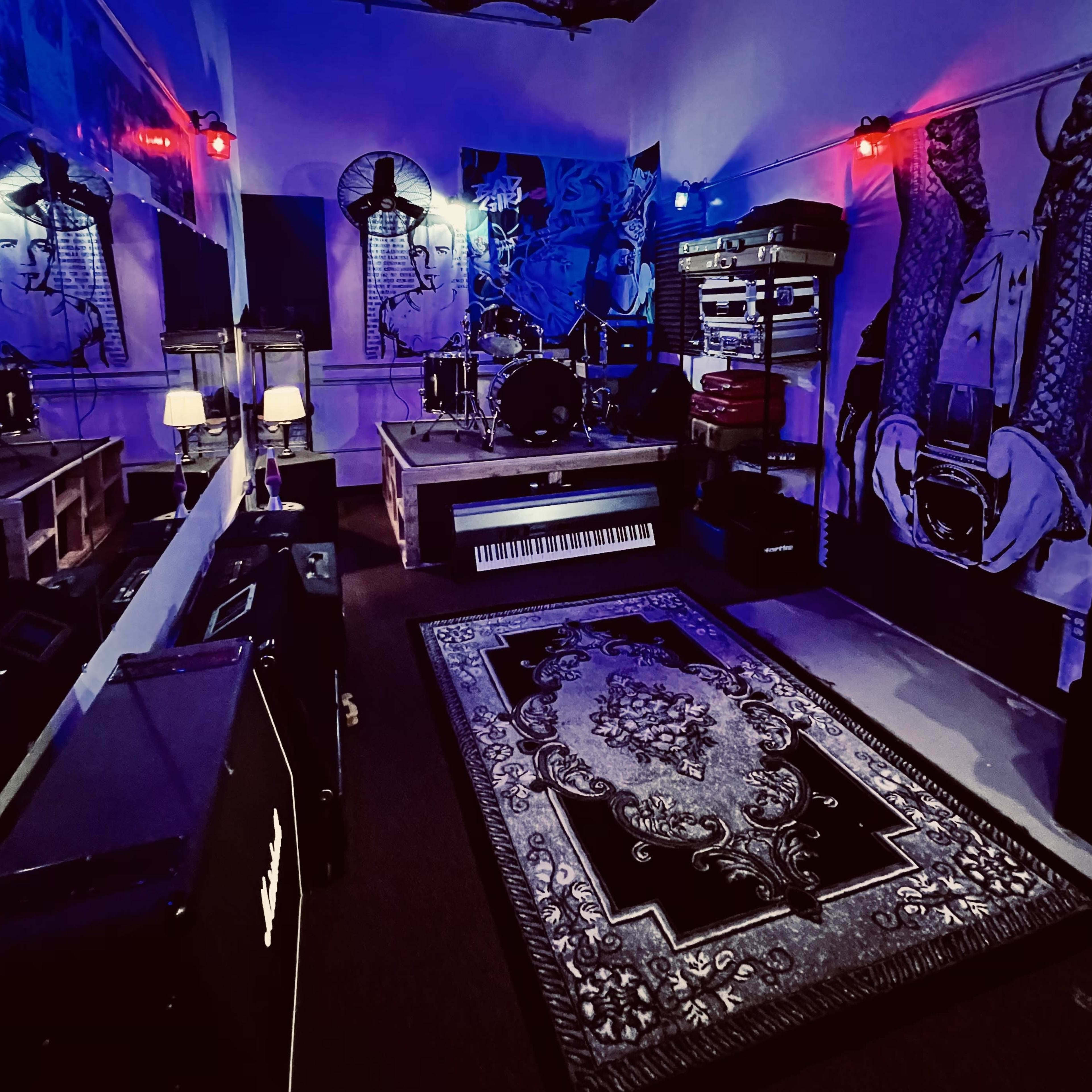 A dimly lit music studio featuring a drum set, keyboards, and amplifiers against a backdrop of artistic murals.