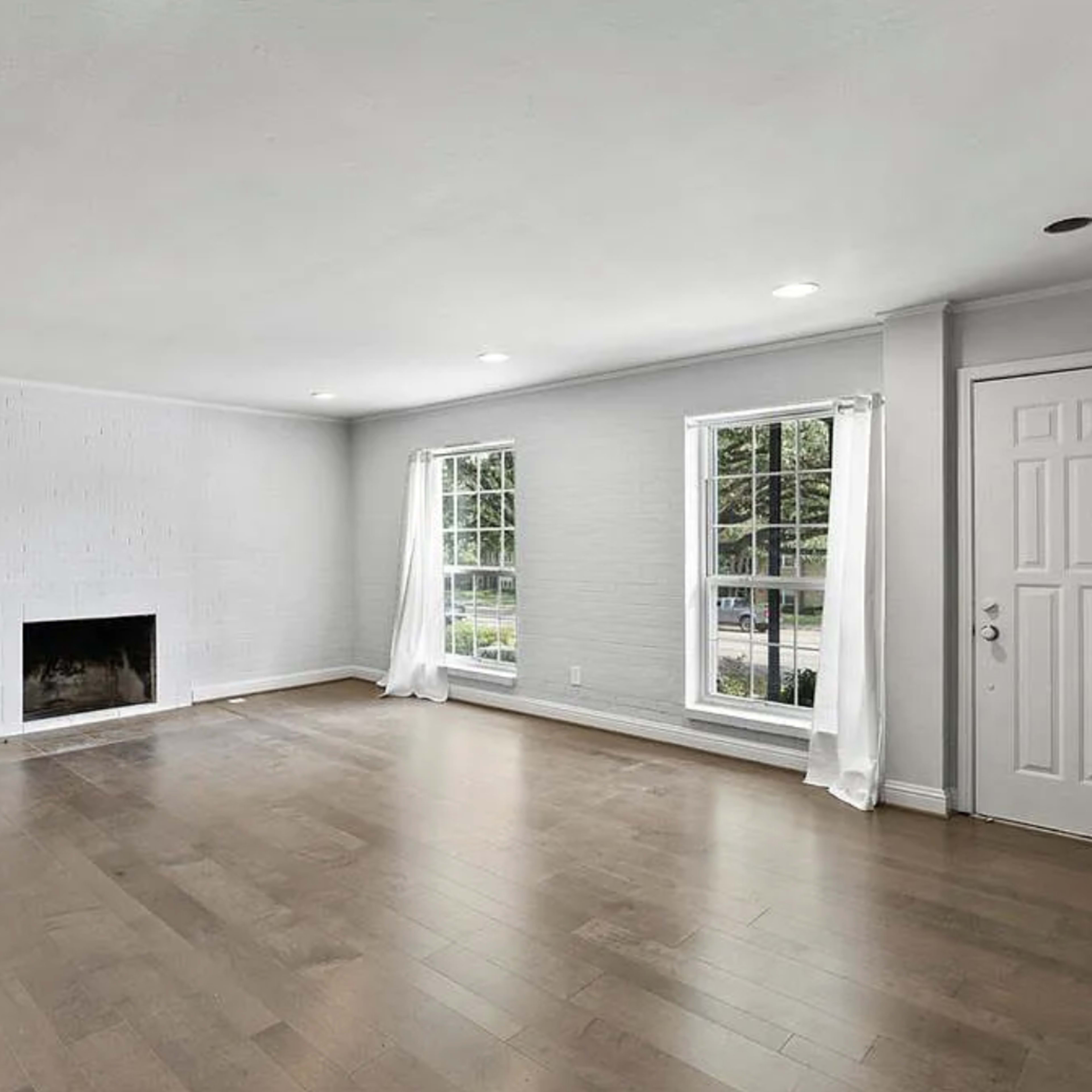 An empty living room with hardwood floors, large windows, and a brick fireplace.