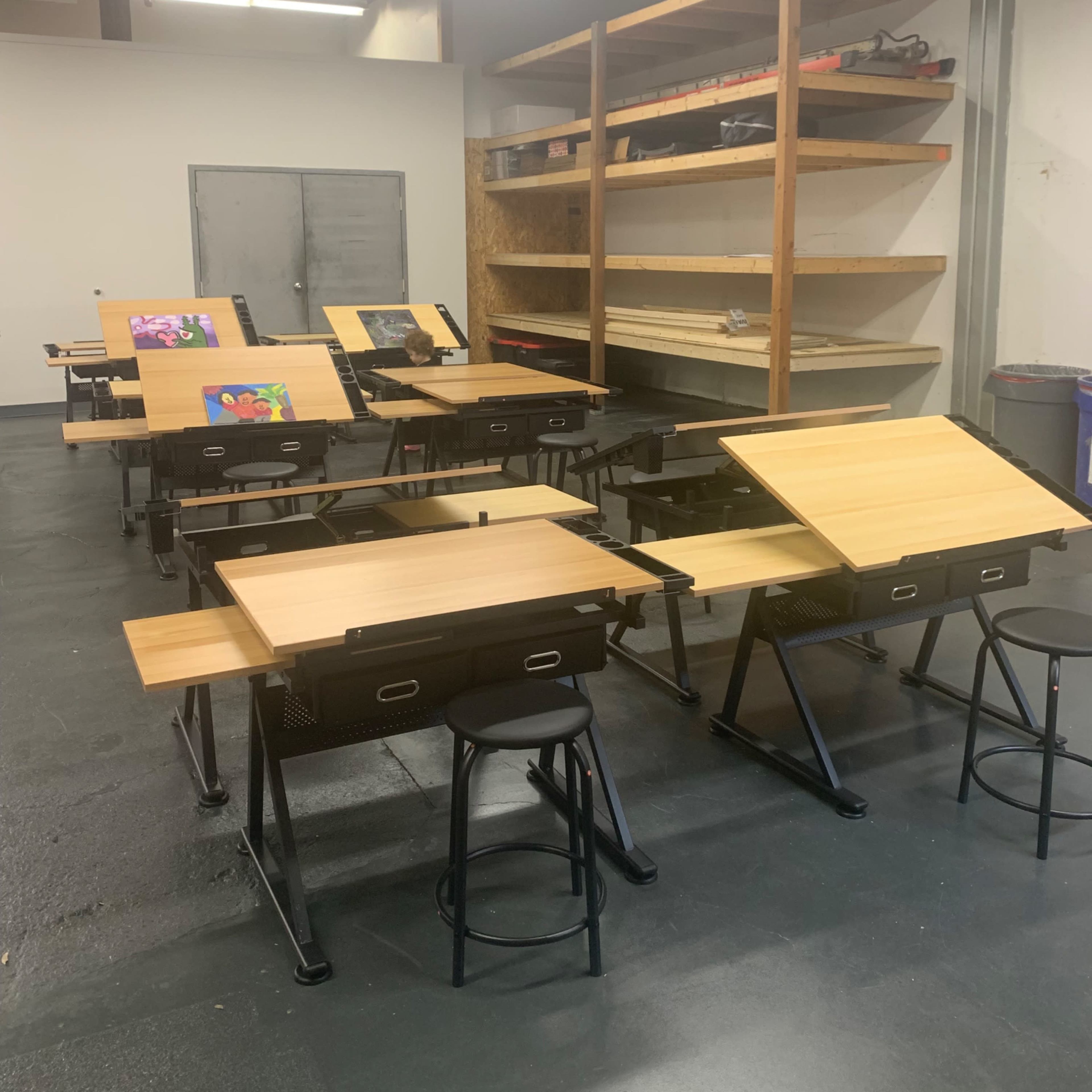A room with multiple drafting tables and stools, arranged in rows, with shelves in the background.