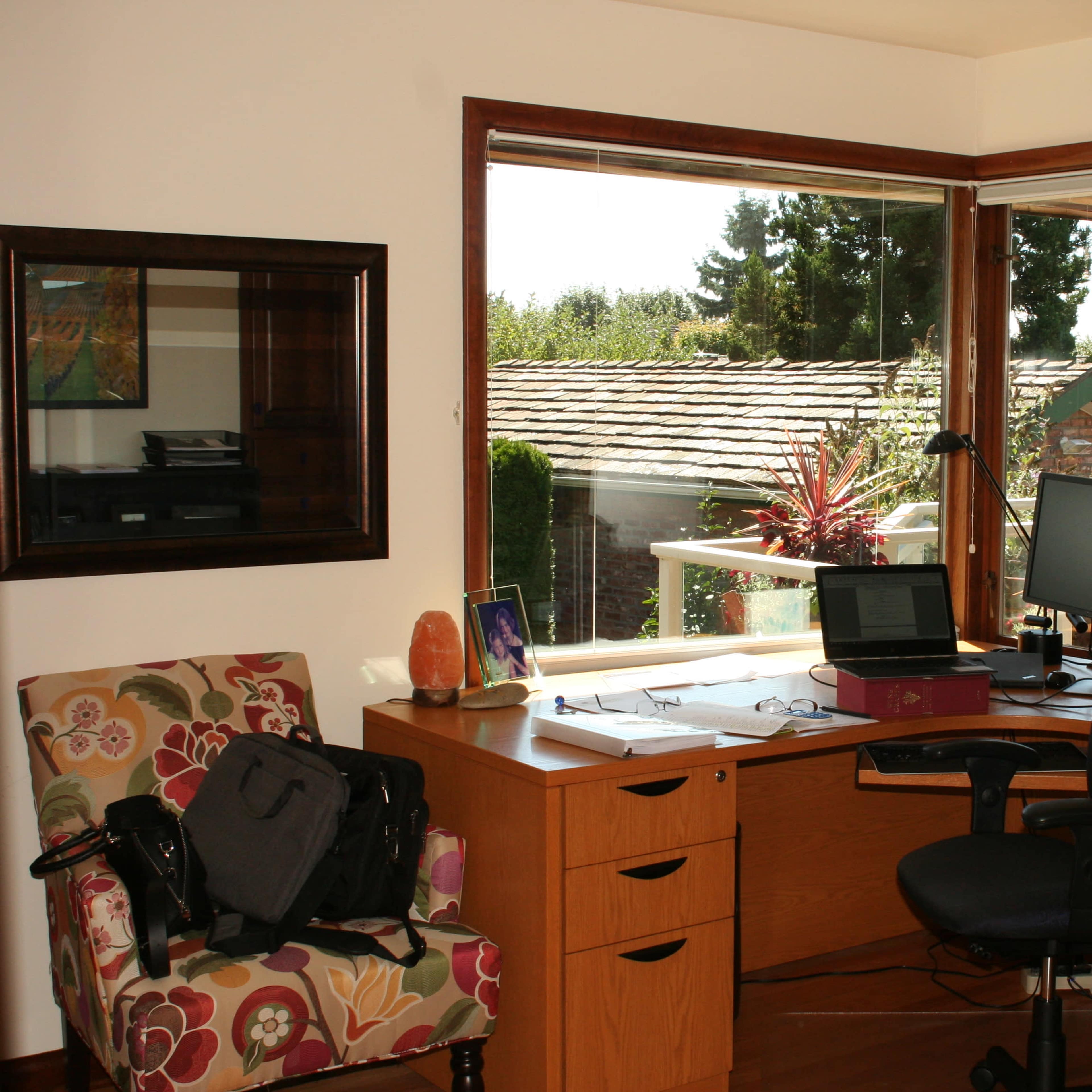 A home office with a wooden desk, a computer setup, and a large window offering a view of greenery outside.