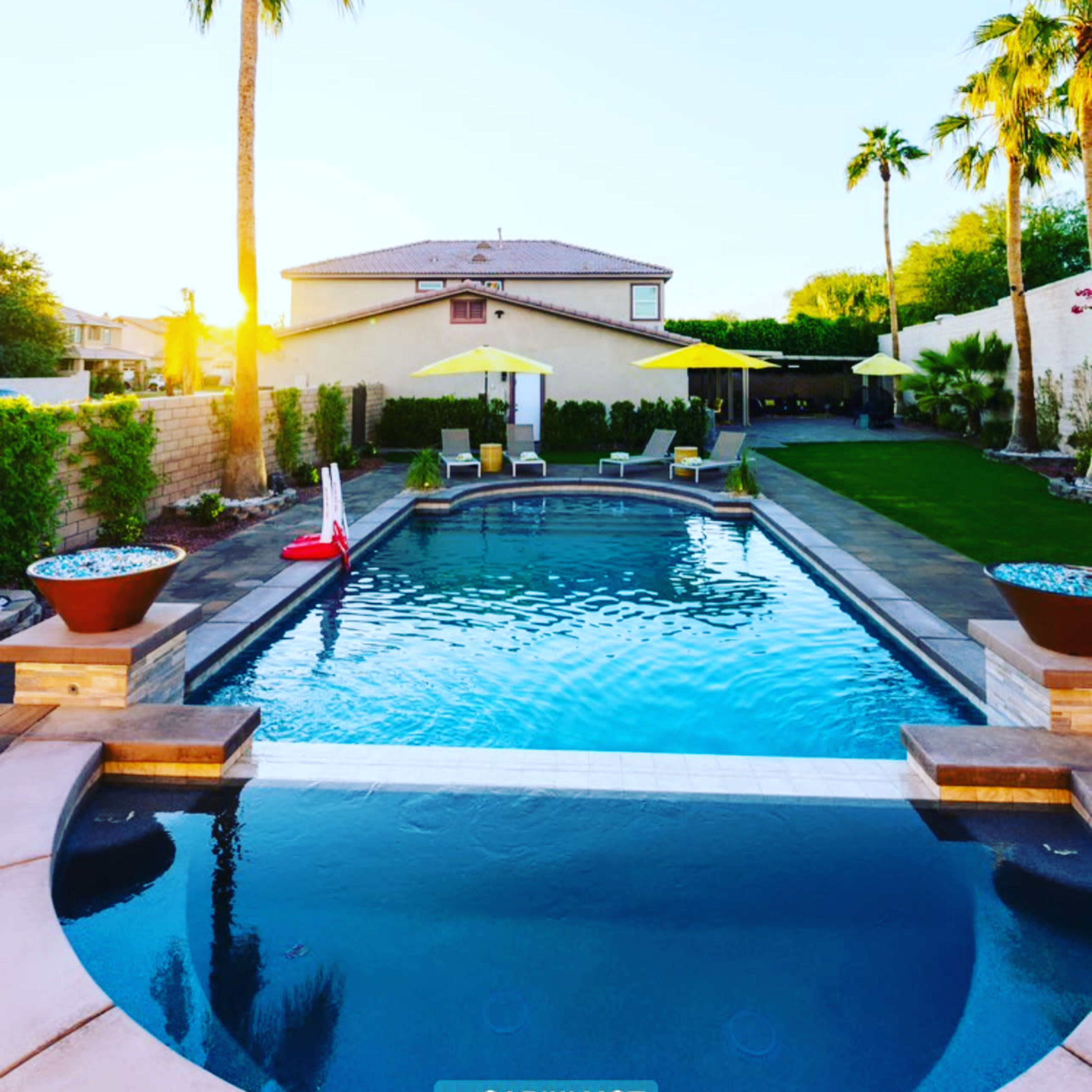 A clear swimming pool surrounded by palm trees and lounge chairs, with a house in the background and a bright sunset.