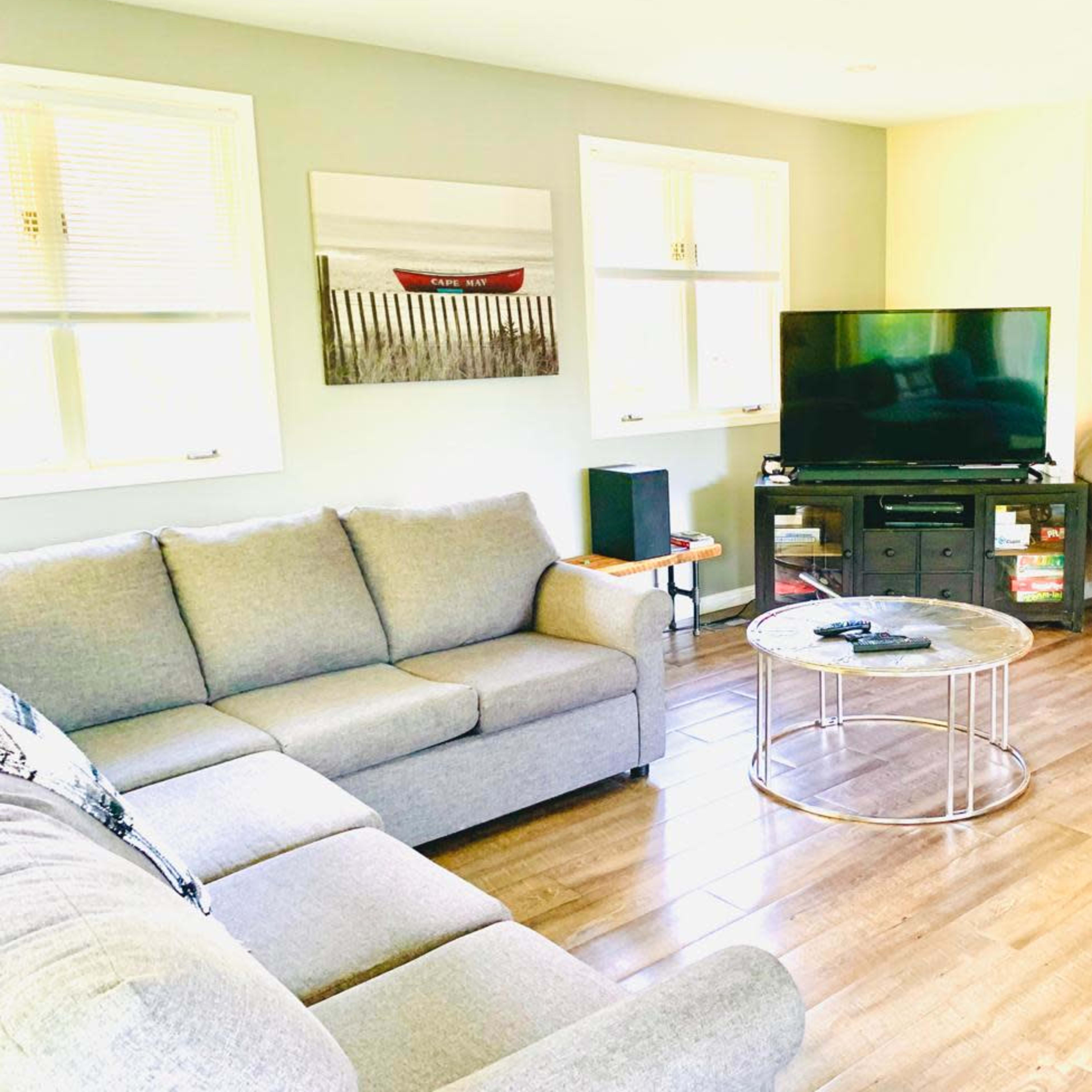 A living room features a gray sectional sofa, a glass coffee table, and a TV on a stand, with light streaming in from windows.