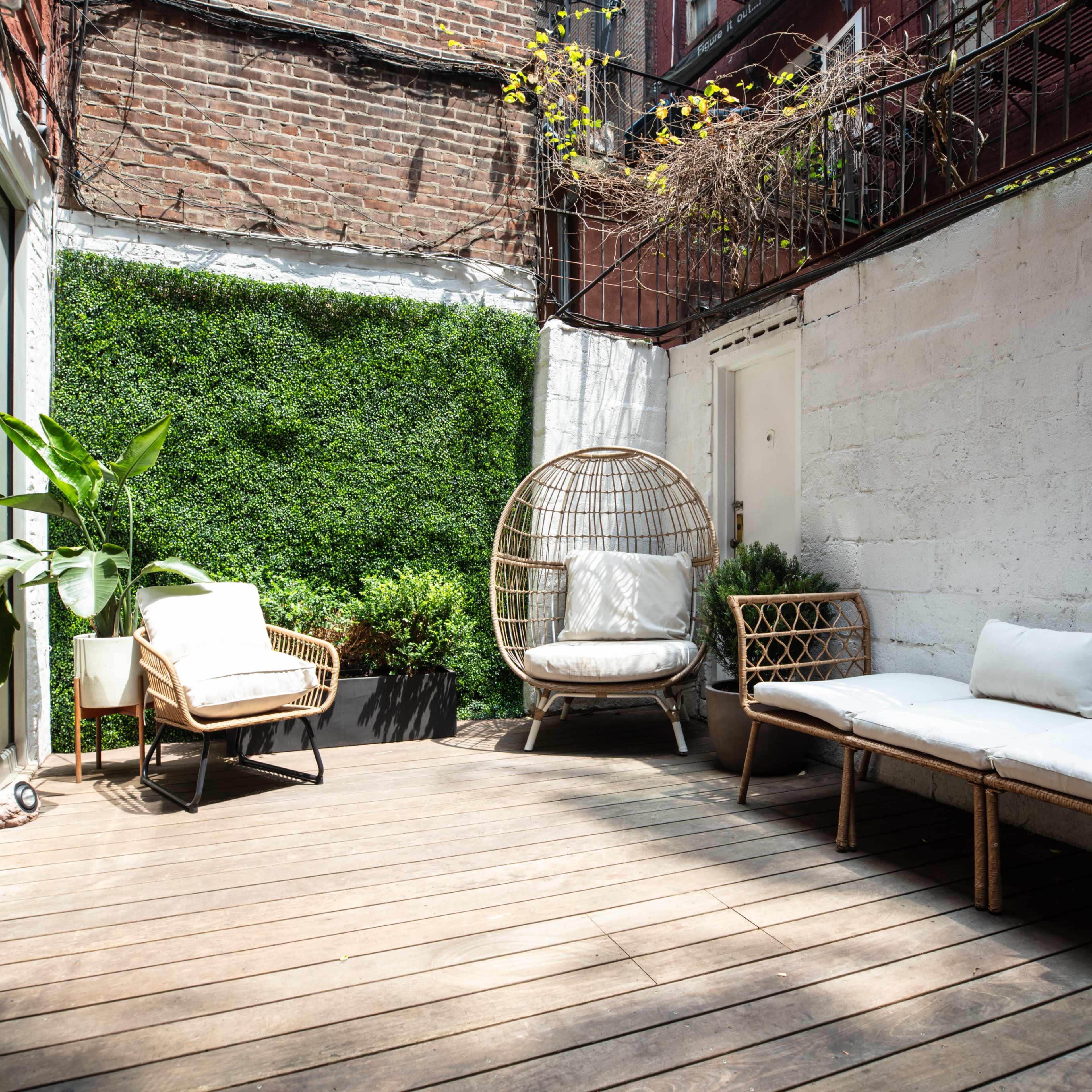 A small outdoor seating area with wooden decking, featuring a hanging wicker chair and several potted plants against a brick wall.