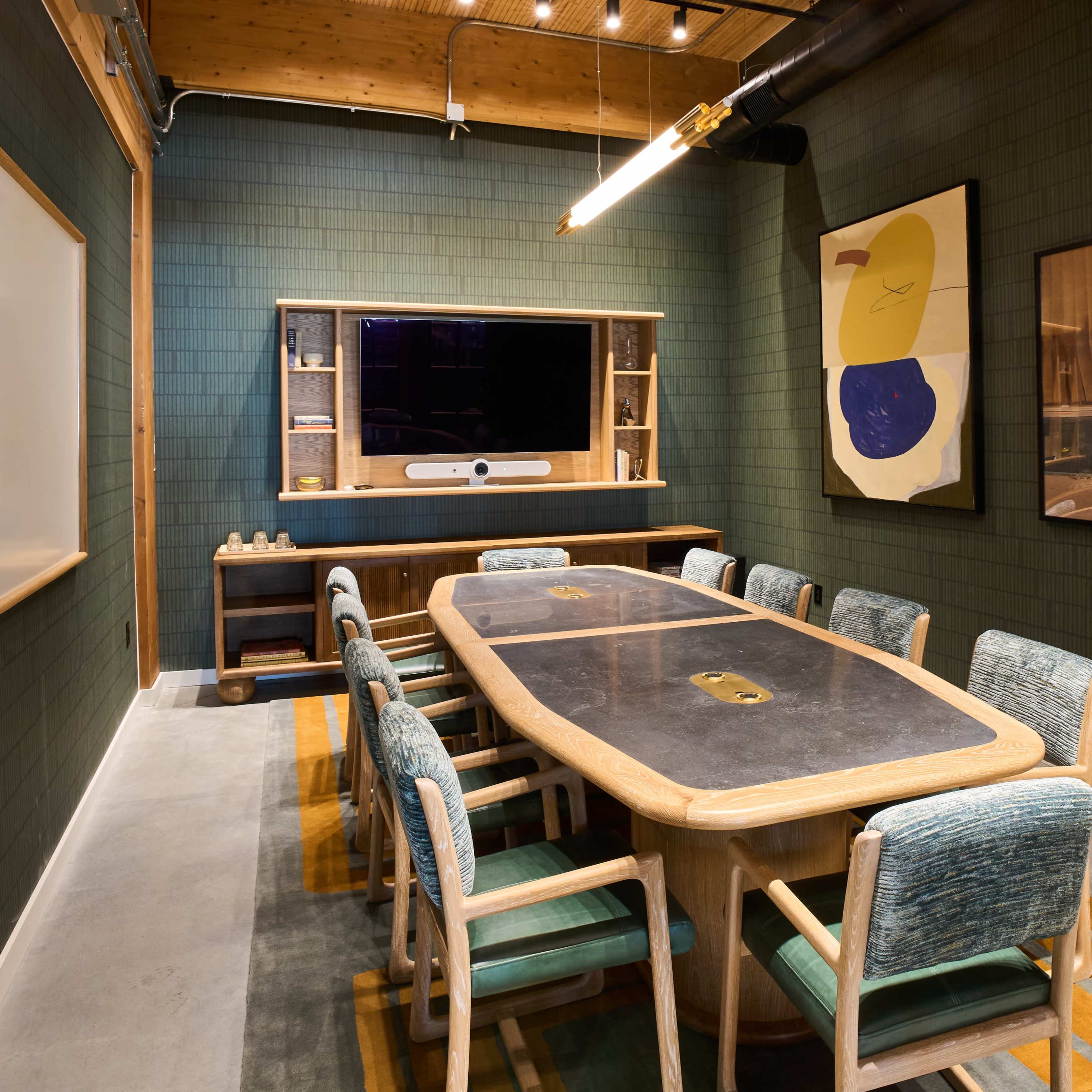 A conference room with a large oval table surrounded by eight chairs, green walls, and a display screen mounted on the back wall.