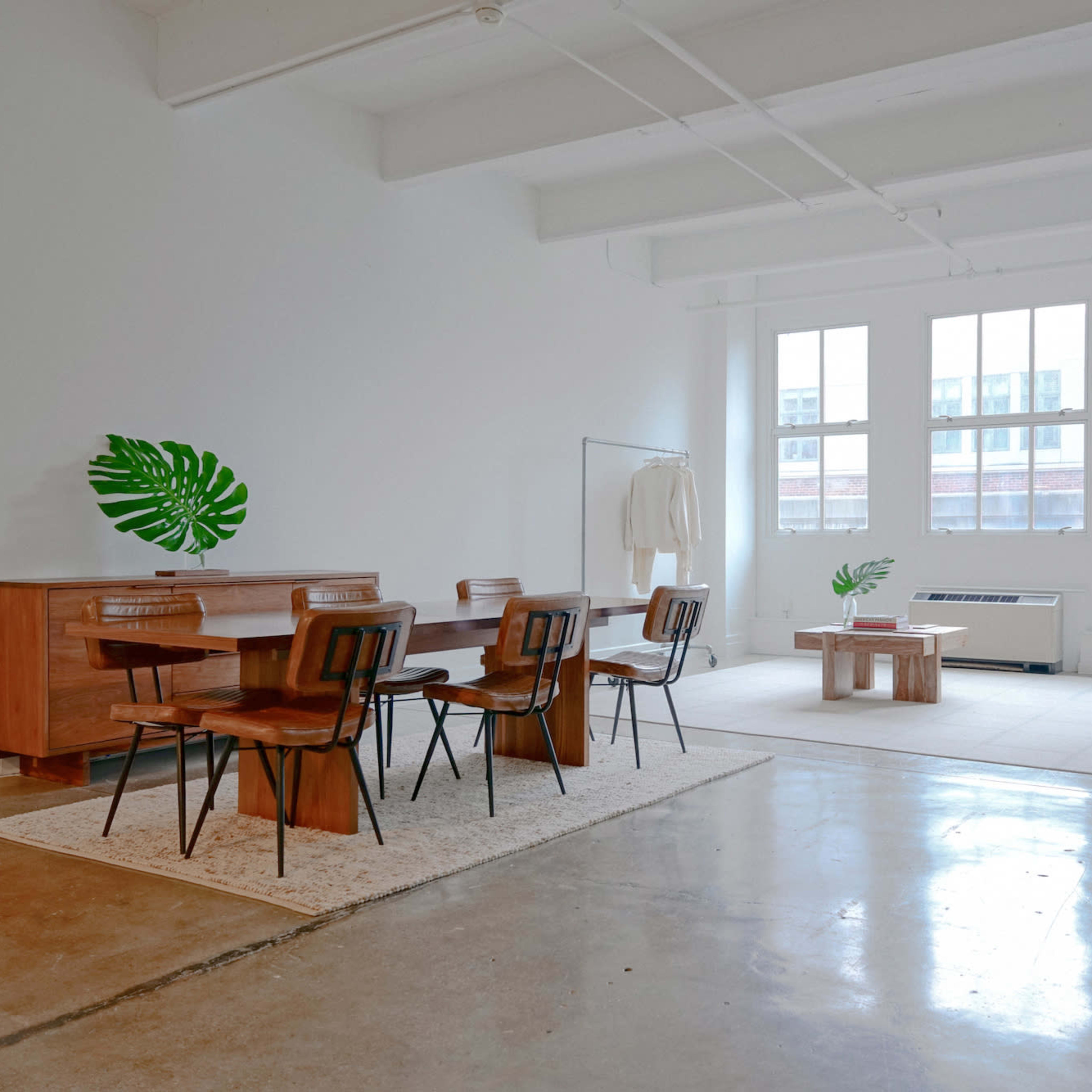 A bright, minimalist interior space featuring a dining area with a wooden table and chairs, and a separate seating area with a small coffee table and plants.