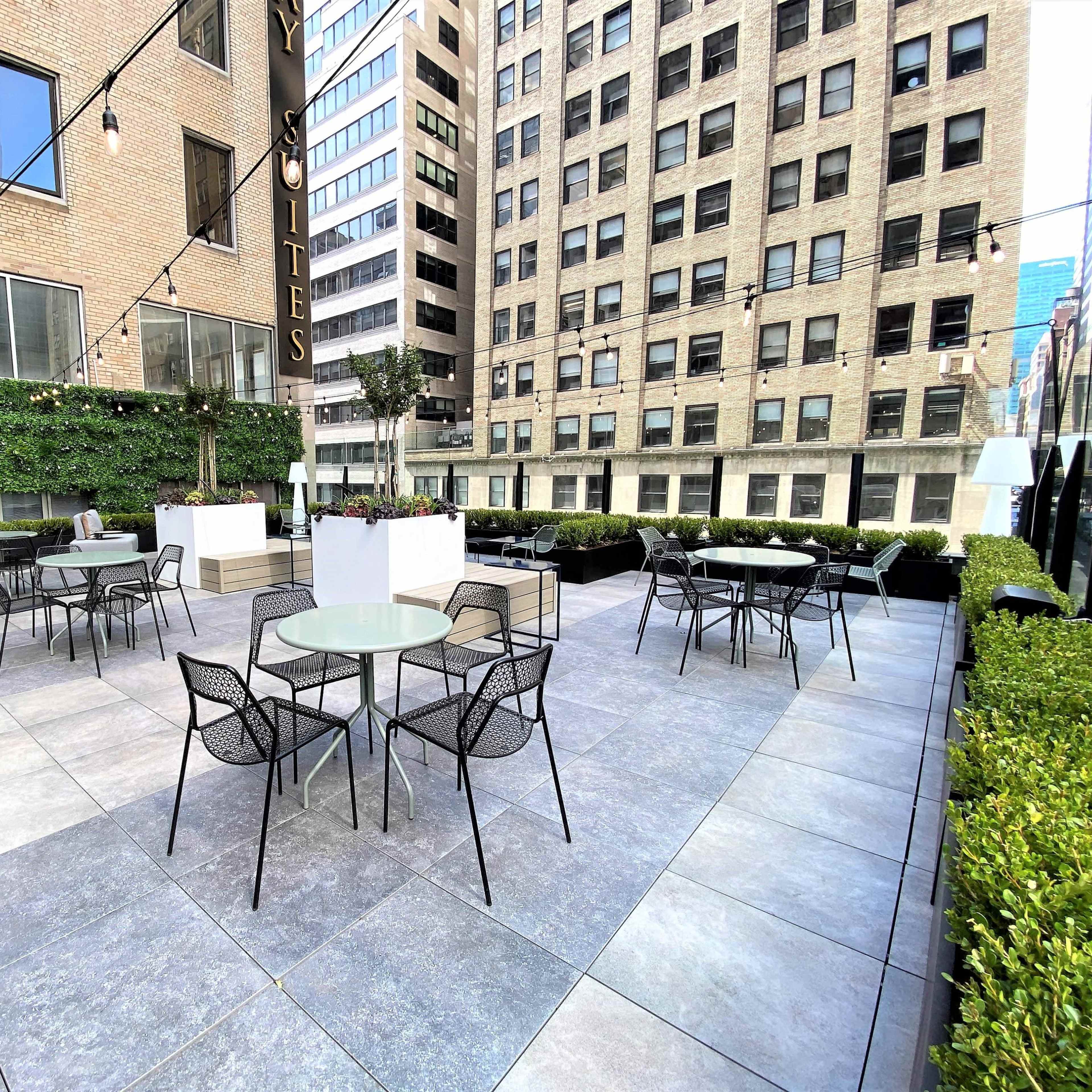 The rooftop patio features several outdoor tables and black chairs surrounded by low hedges and tall buildings.
