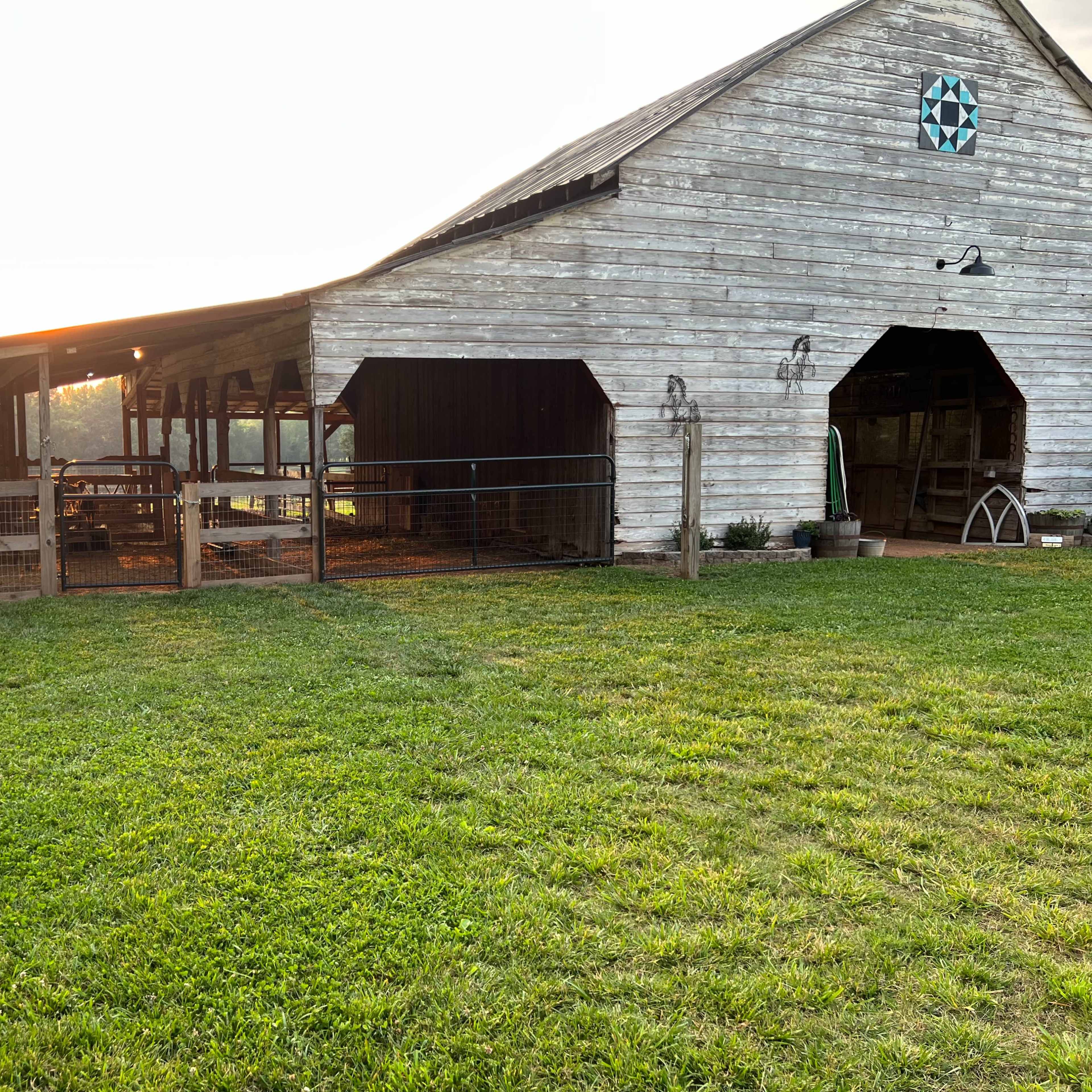 A weathered barn sits on a grassy field, with sunlight illuminating one side and an adjoining fenced area.